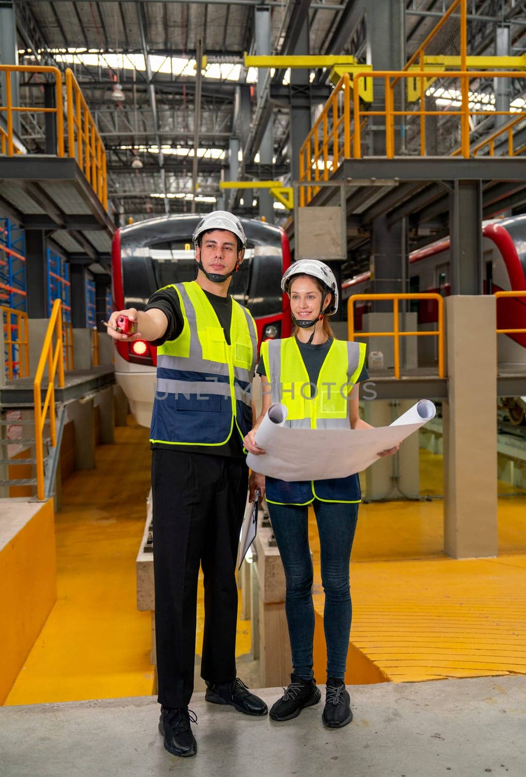Verital image of two professional engineer or technician workers man and woman discuss with drawing paper and the man point to left side also stay in front of electric train in factory workplace.