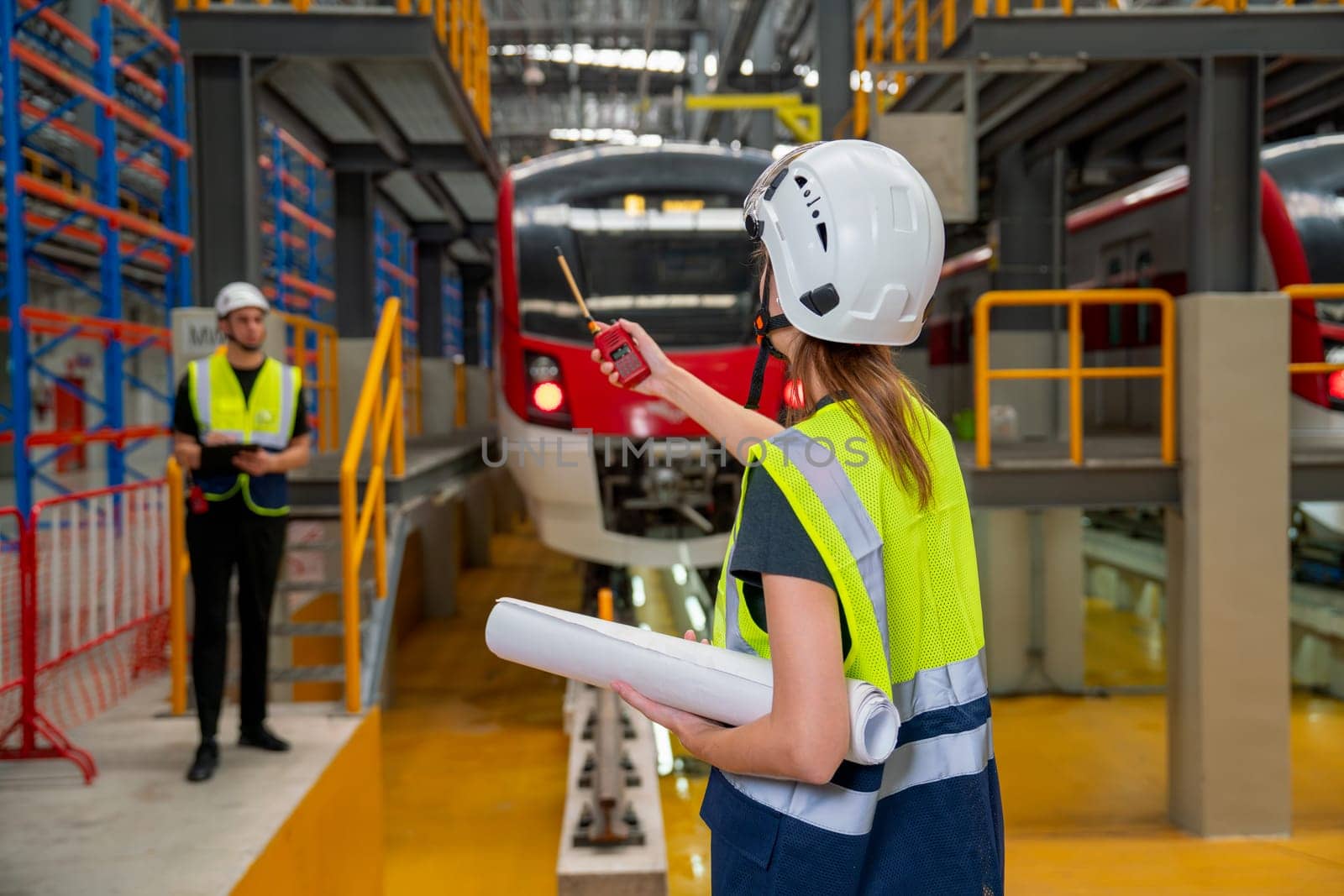 Back of technician or engineer worker woman use walkie talkie point to her co-worker during work in front of electric train in factory workplace or maintenance center.
