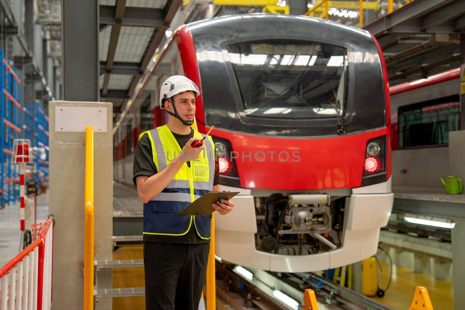 Technician or engineer worker man hold walkie talkie to contact with team member or staff also stay in front of electric train in factory workplace or maintenance center.