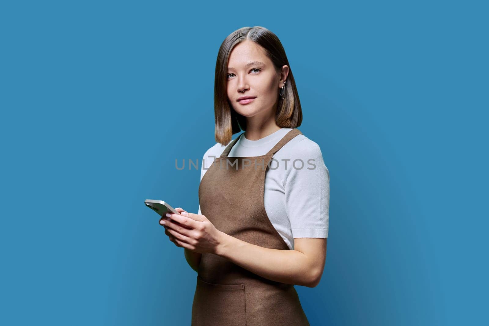 Portrait of young woman in apron holding smartphone in hands on blue background. Serious female using mobile phone, looking at camera. Technologies, applications apps, service, small business, work
