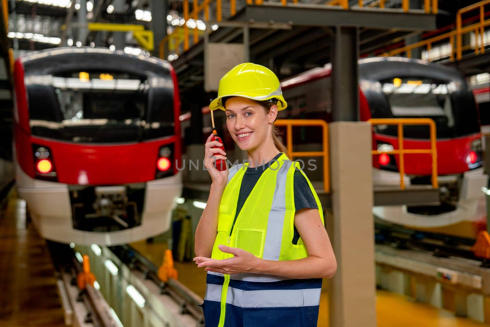 Engineer or technician worker woman hold walkie talkie and look at camera also stay in front of electric train in factory workplace or maintenance center.