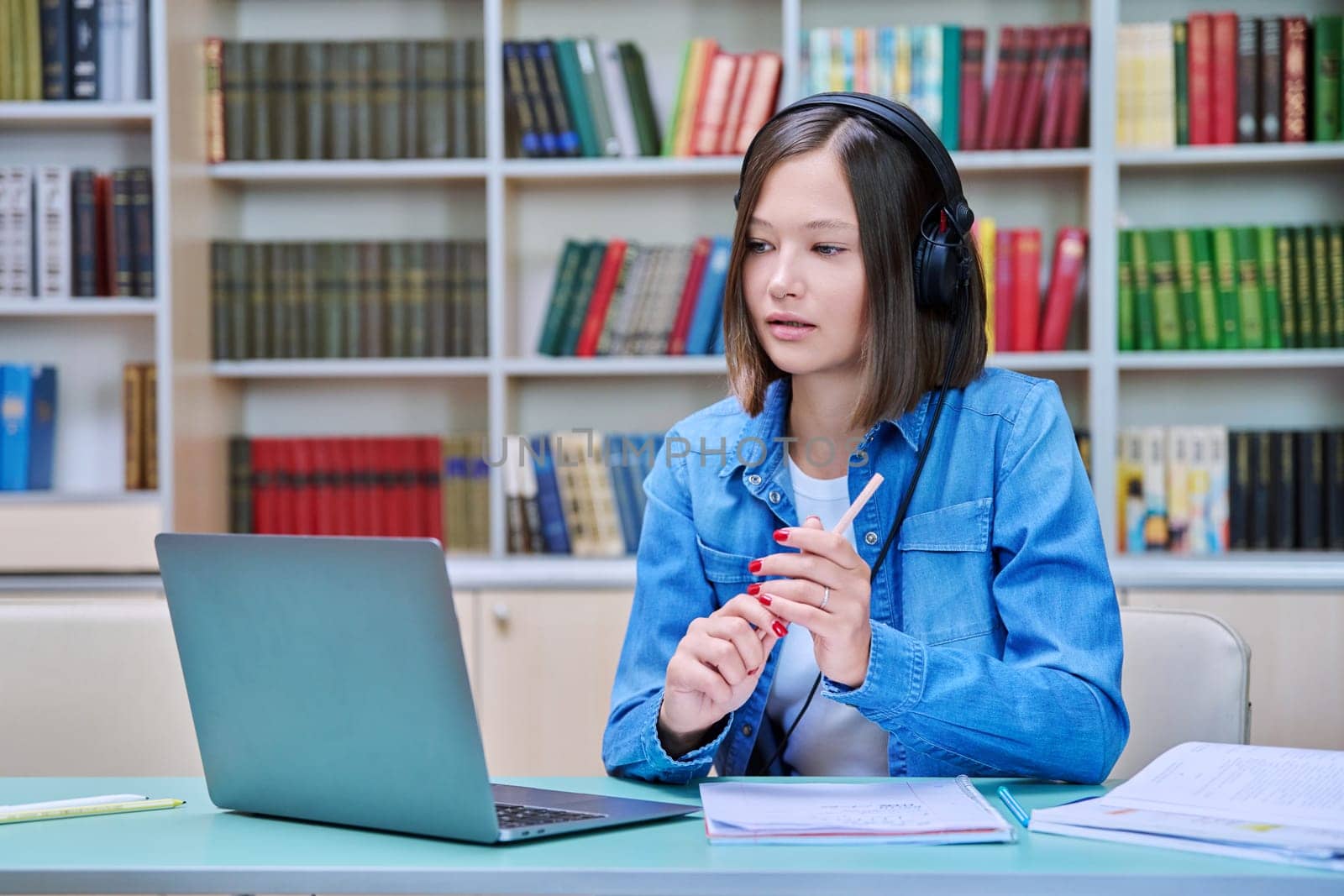 Young female college student in headphones sitting at desk with laptop having online video conference chat, looking at screen talking listening watching learning class, webinar, lesson inside library