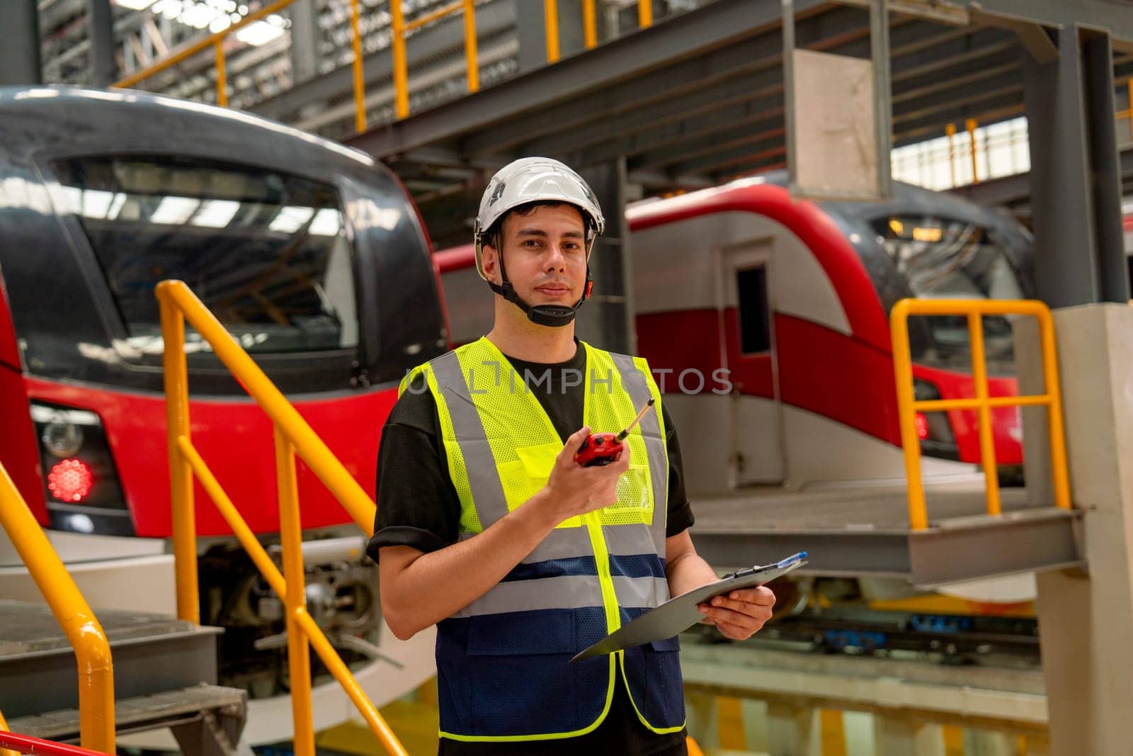 Portrait of engineer or technician worker man hold walkie talkie and archboard also look at camera with stand in front of electric train in factory workplace or maintenance center.