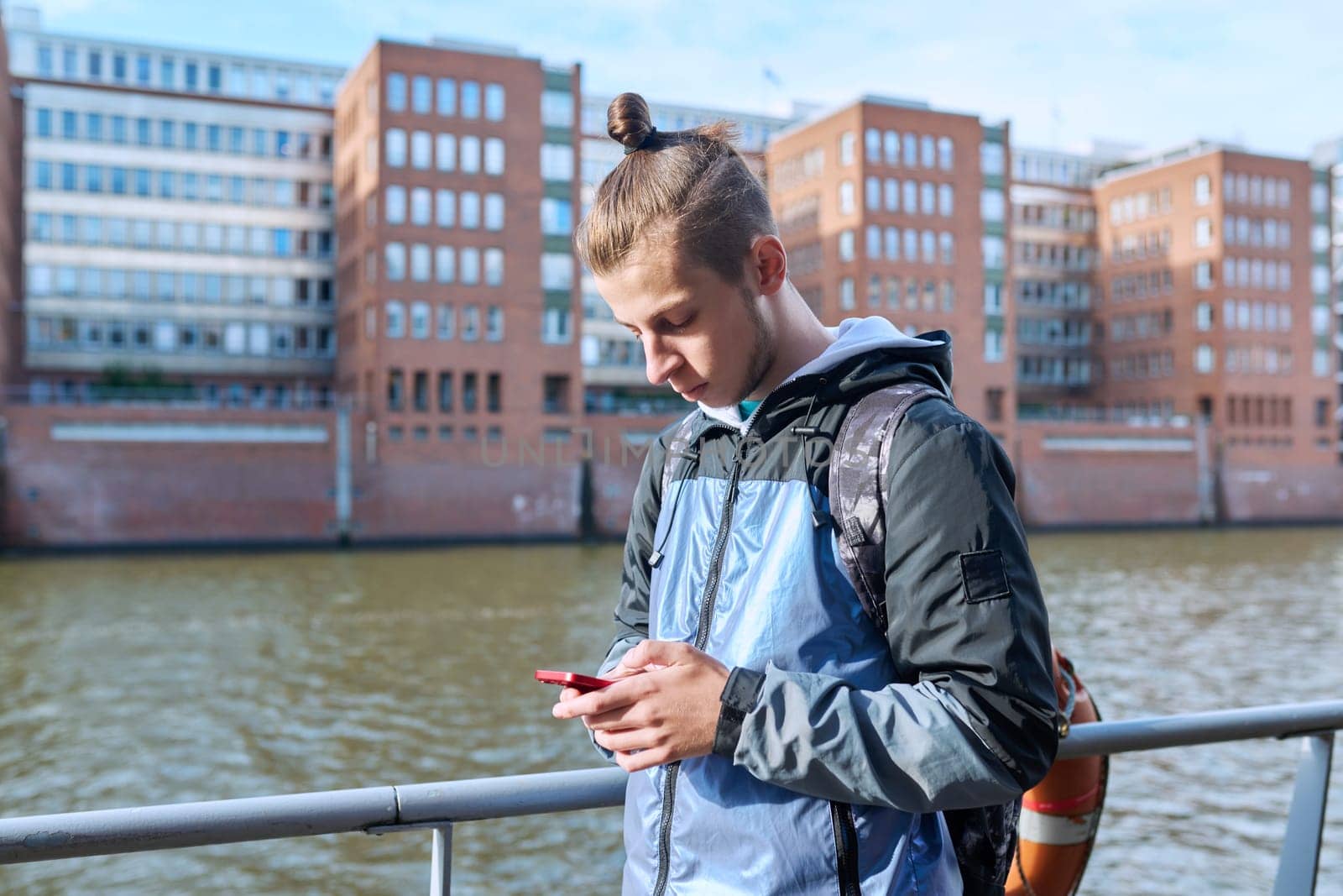 Fashionable young male with backpack using smartphone outdoors in city. Hipster with trendy hairstyle beard with phone in hands mobile applications for tourism travel communication entertainment study