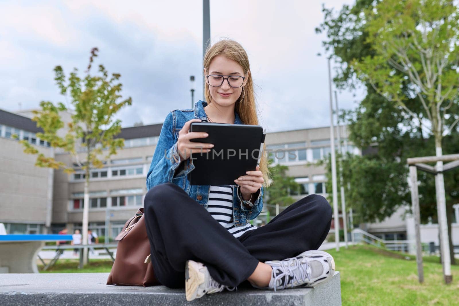 Girl student teenager outdoor near school building. Smiling teenage female with backpack, looking at screen of digital tablet. Adolescence, education, learning concept