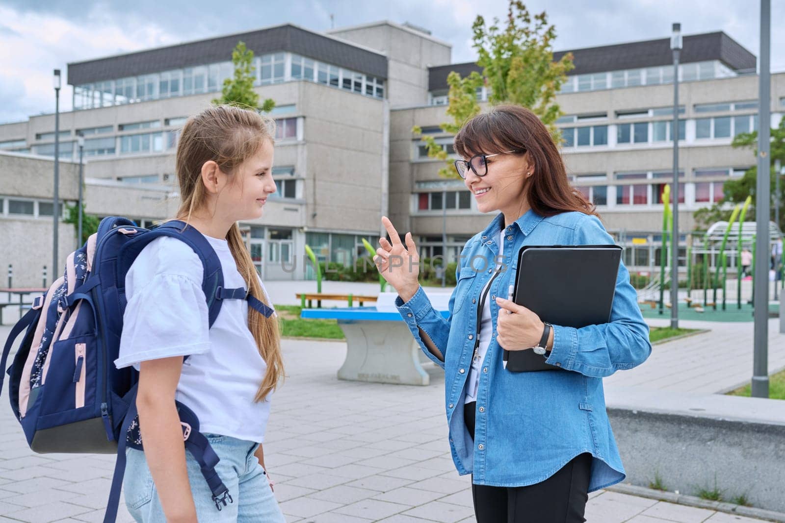 Talking female teacher and schoolgirl child outdoor, school building background. Meeting communication student girl with backpack and mentor counselor. Education, pre-teenage, learning, back to school