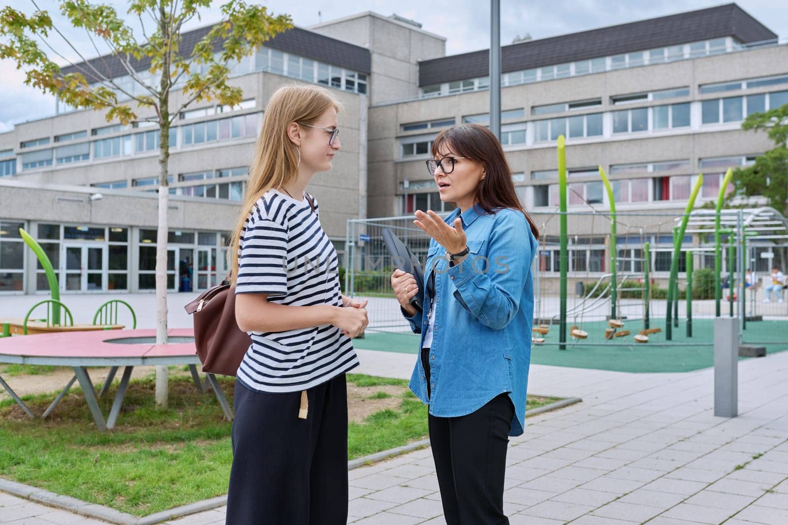 Talking female teacher and teenage high schoolgirl outdoor, school building background. Meeting communication student girl with backpack and mentor counselor. Education, adolescence, learning concept