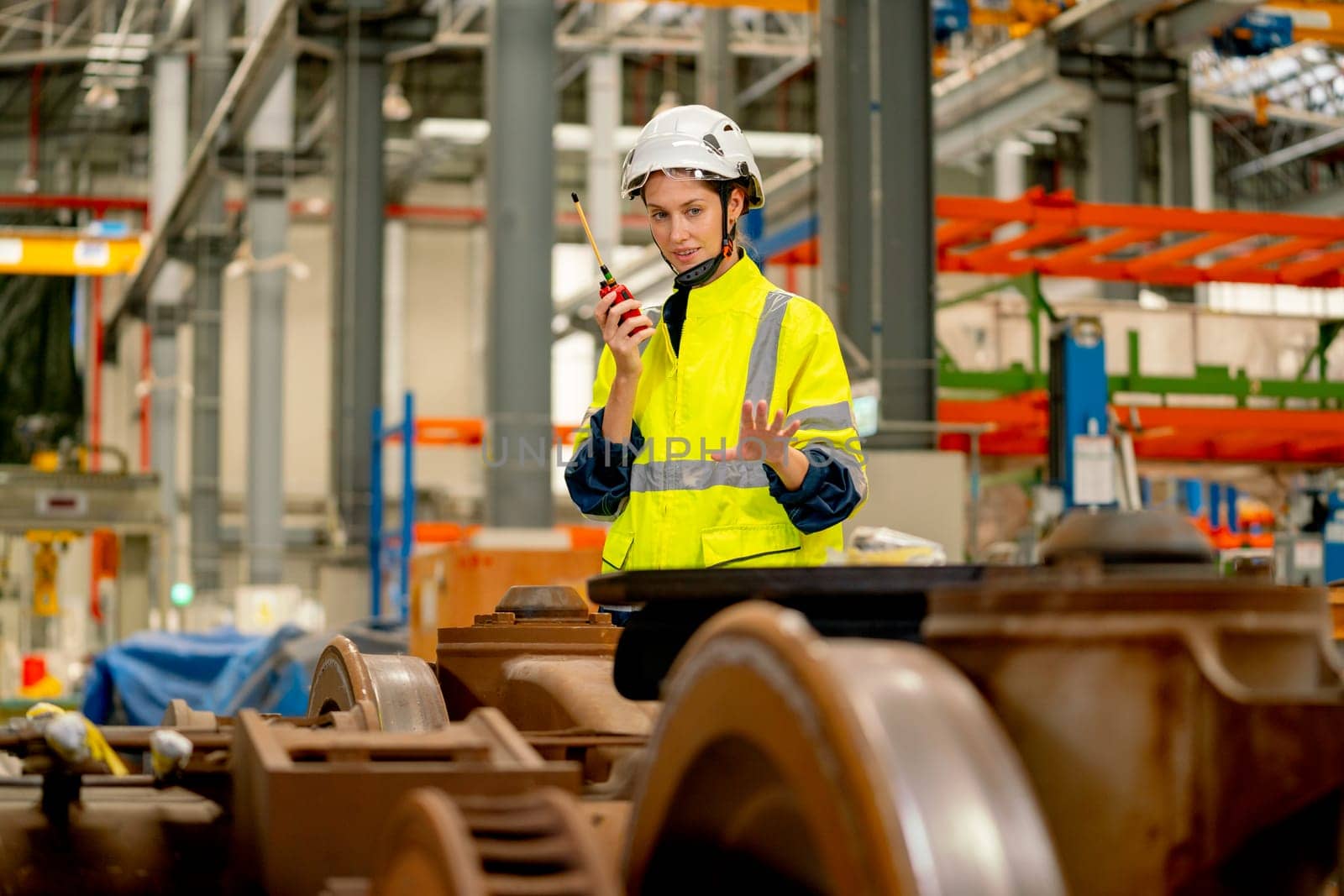 Profession engineer or technician worker use walkie talkie to contact with co-worker or team staff and look to wheel and stand behind part of electric train in maintenance center.