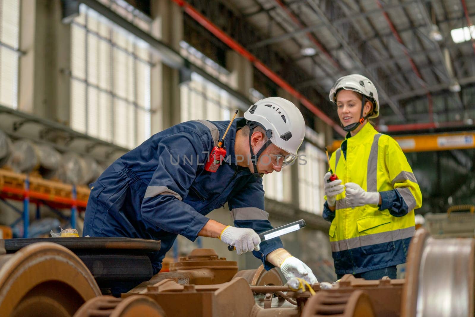 Professional technician man hold light stick to check and fix problem of parts of electric train and support by factory worker woman who stand beside in workplace area.