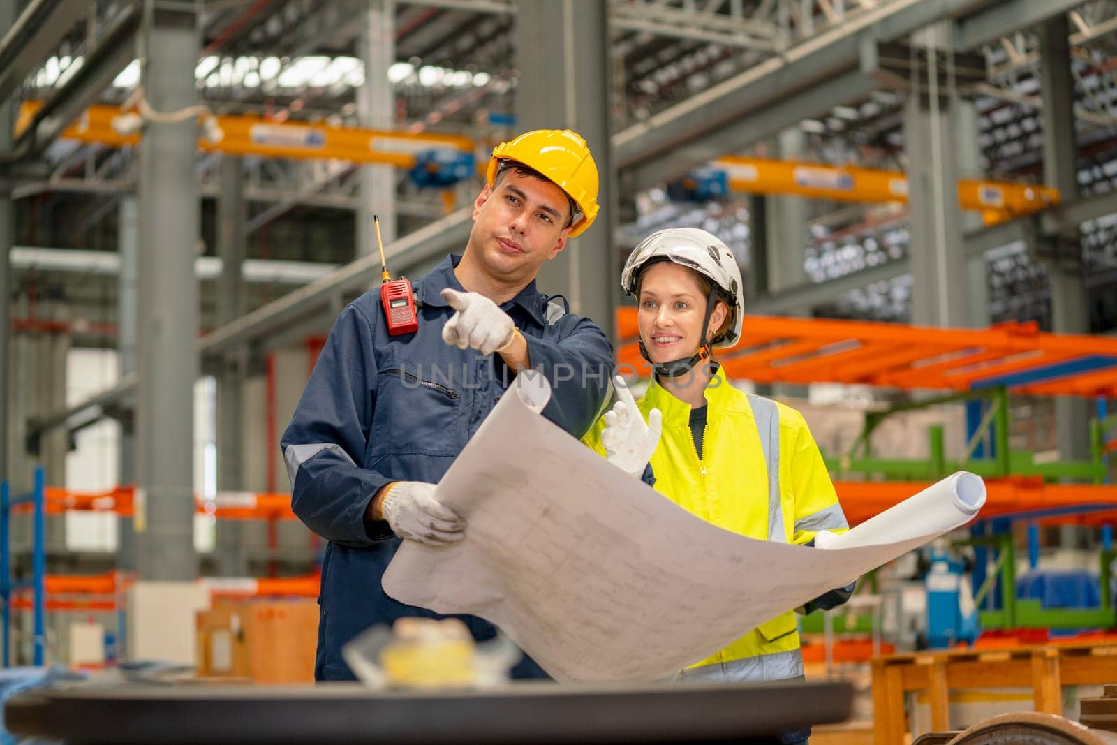 Factory workers man and woman discuss about the project using drawing paper or plan in workplace with machine and product shelves are in the background.