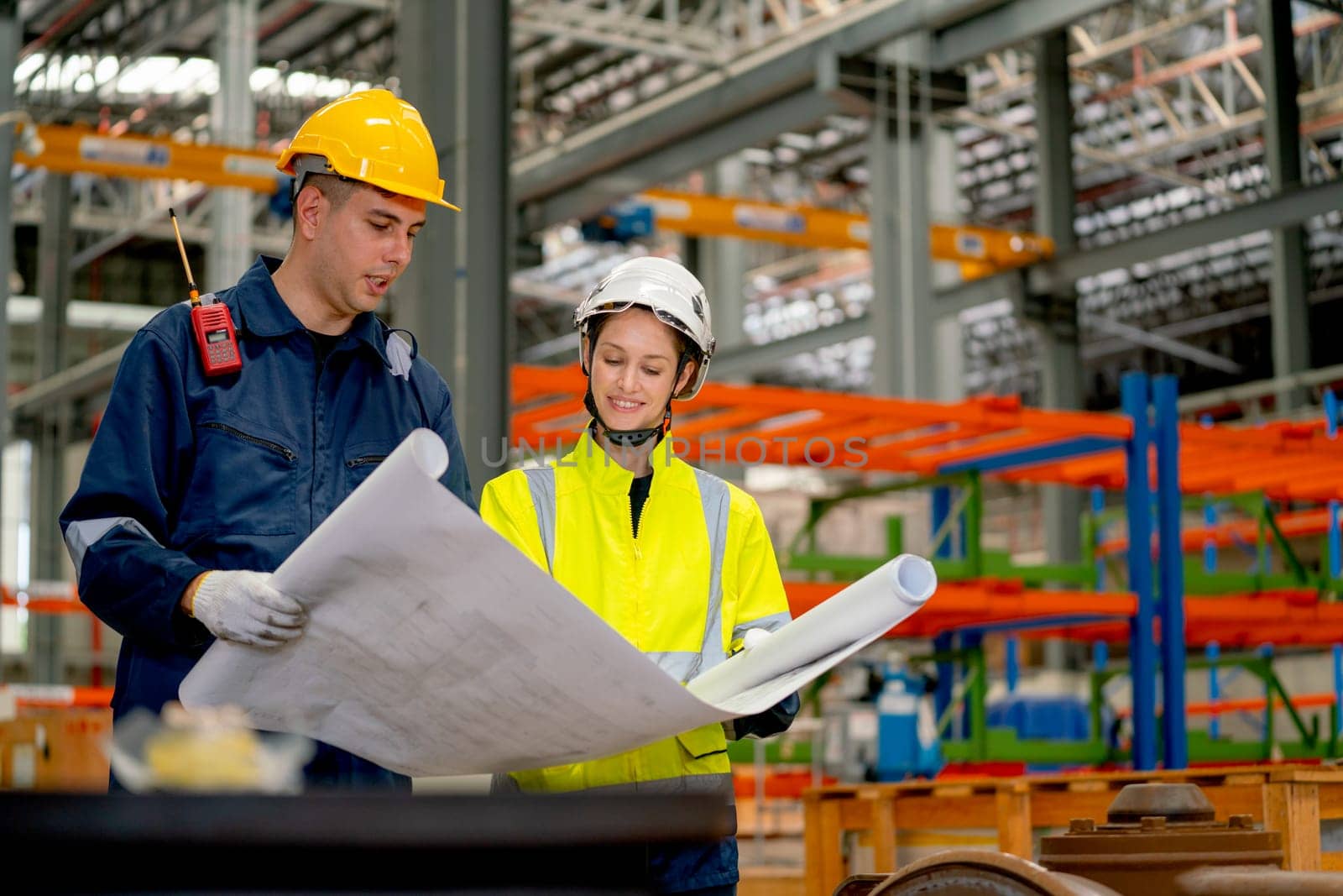 Factory workers man and woman discuss about the project using drawing paper or plan in workplace with machine and product shelves are in the background.