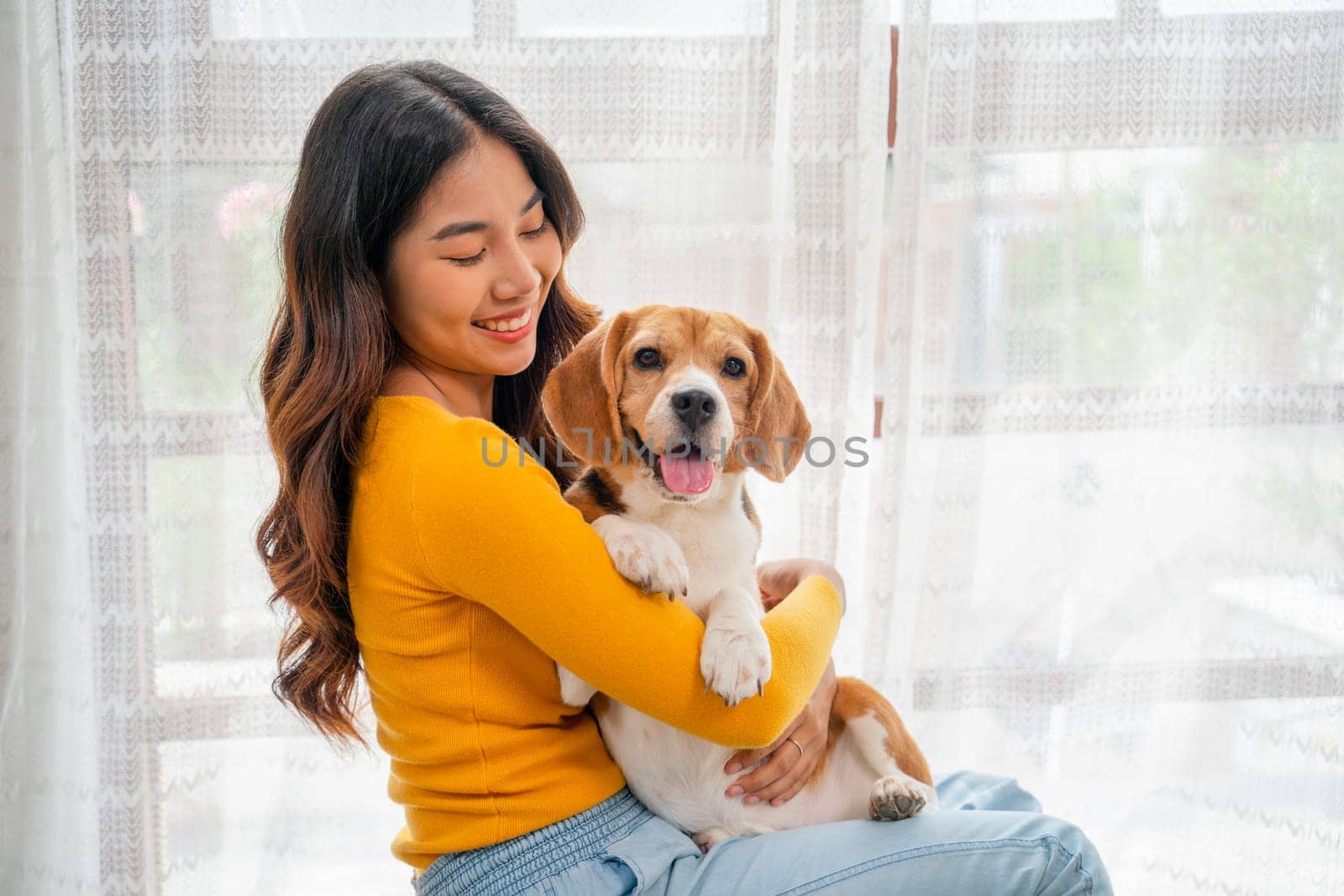 Close up young Asian girl hold and hug beagle dog and sit in front of glass door in her house and she look happy to play fun together.