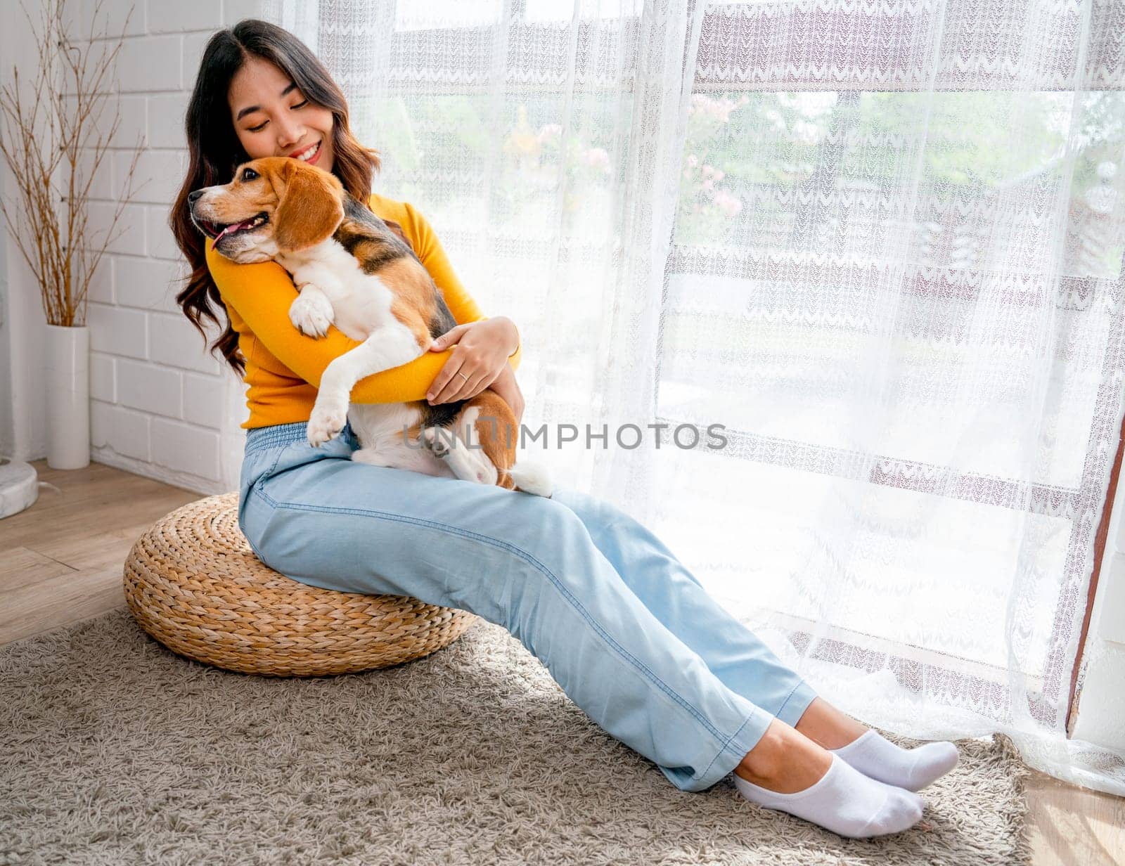 Young Asian girl hold and hug beagle dog and sit in front of glass door in her house and she look happy to play fun together. by nrradmin