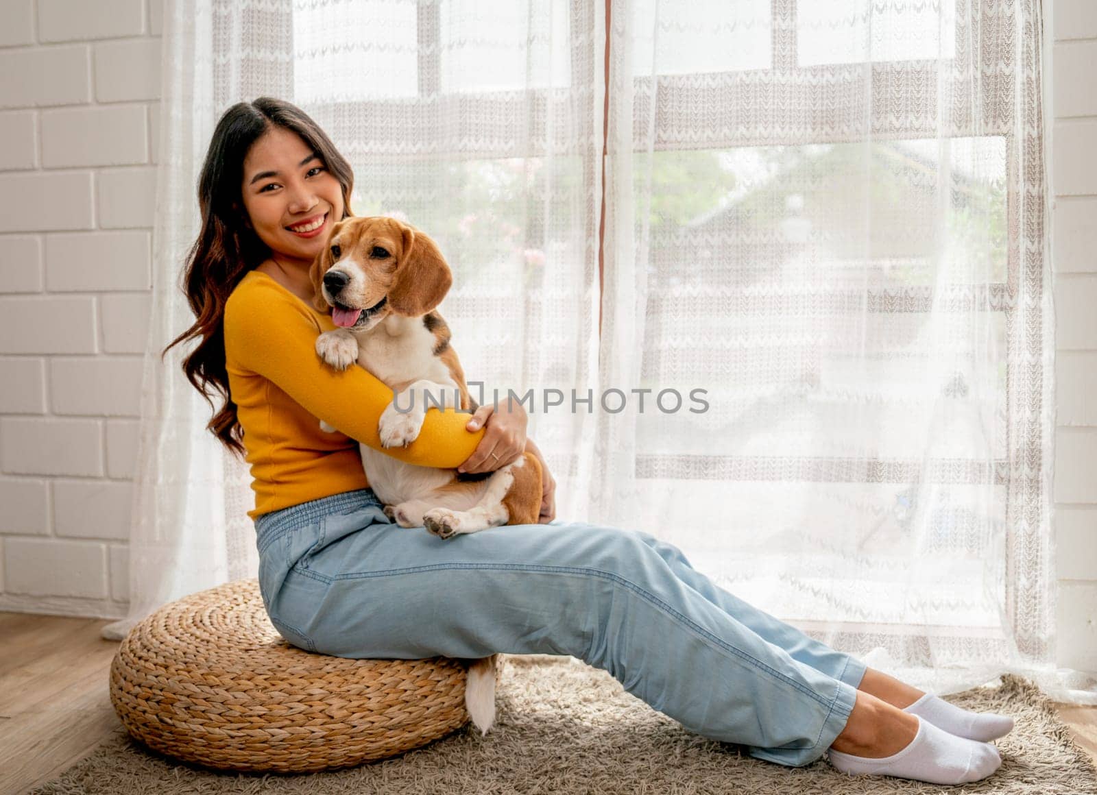 Young Asian girl look at camera with smiling also hold and hug beagle dog and sit in front of glass door in her house and she look happy to play fun together. by nrradmin