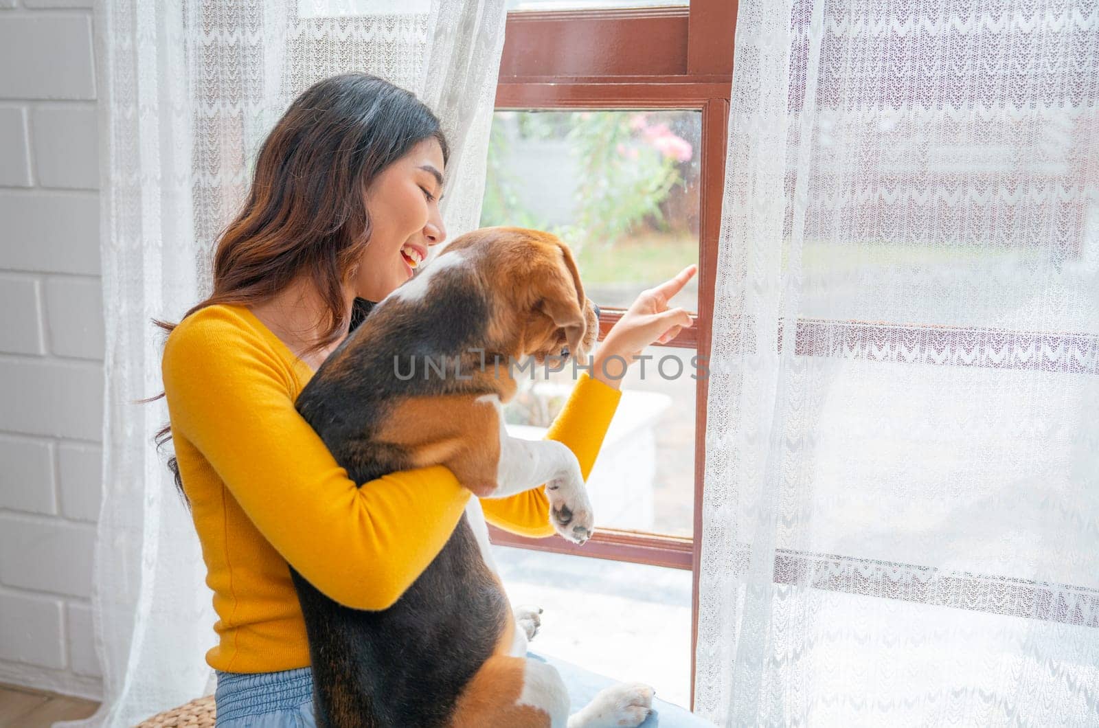 Young Asian woman hold and hug beagle dog also point outside through glass windows to relax and fun with her pet in the house.