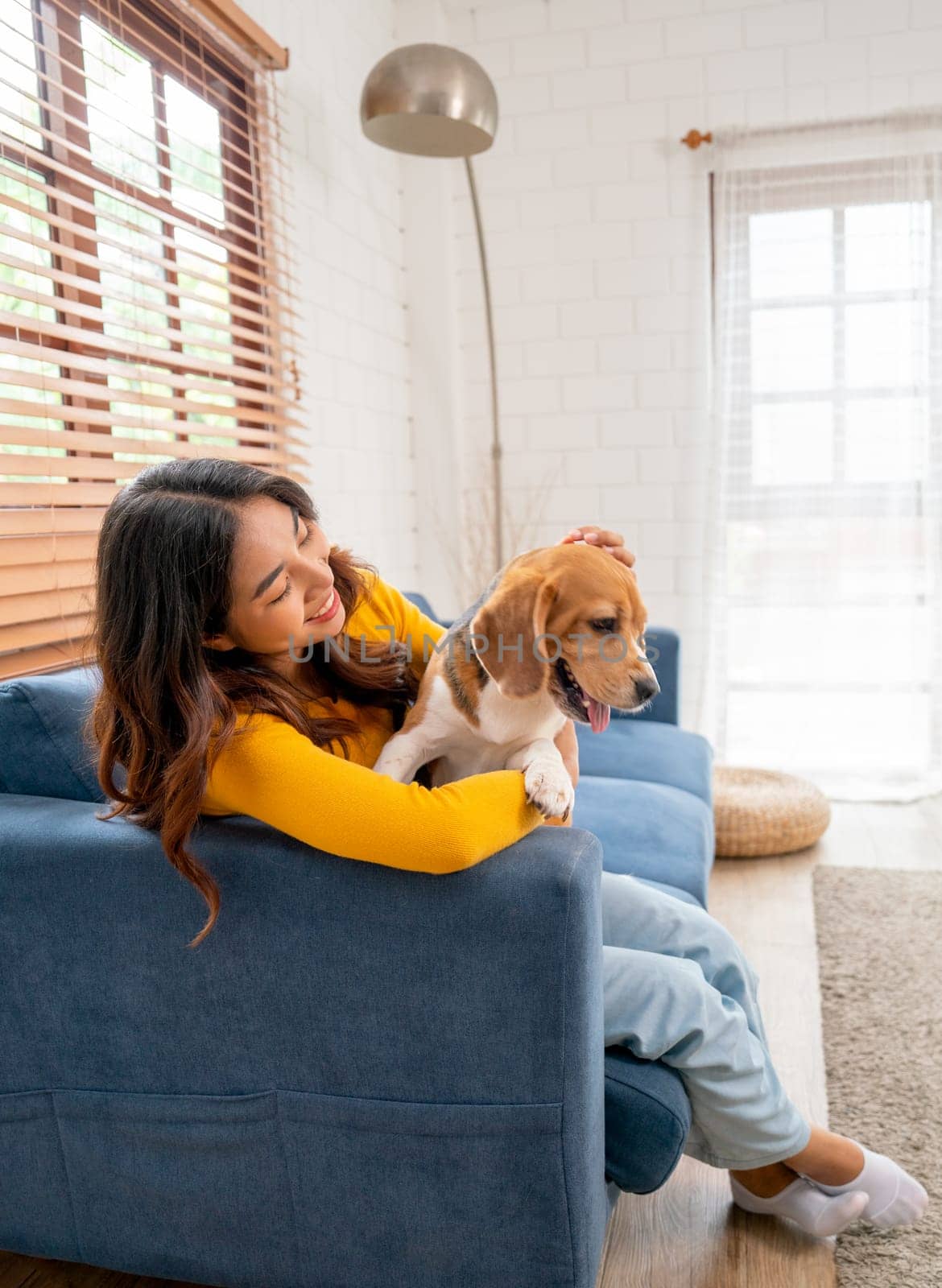 Vertical image of young Asian girl hug beagle dog and stay on sofa of living room of her house with day light. by nrradmin