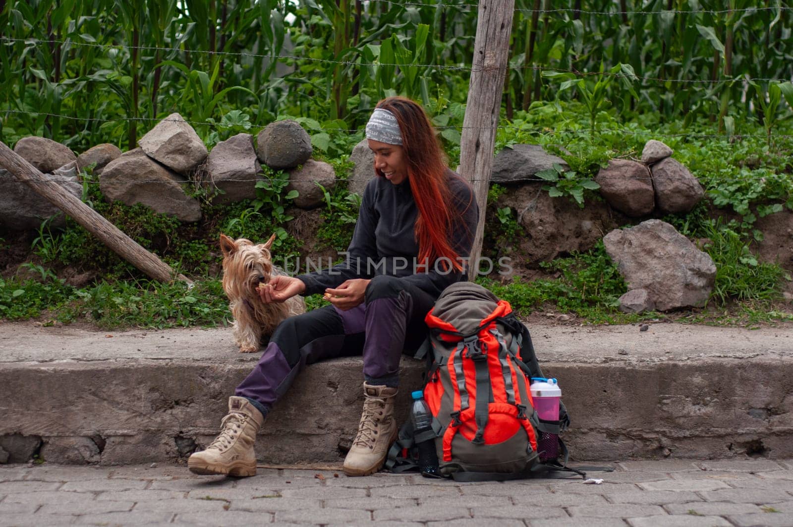 woman backpacker sitting on the sidewalk feeding her little dog some cookies and an orange backpack. by Raulmartin