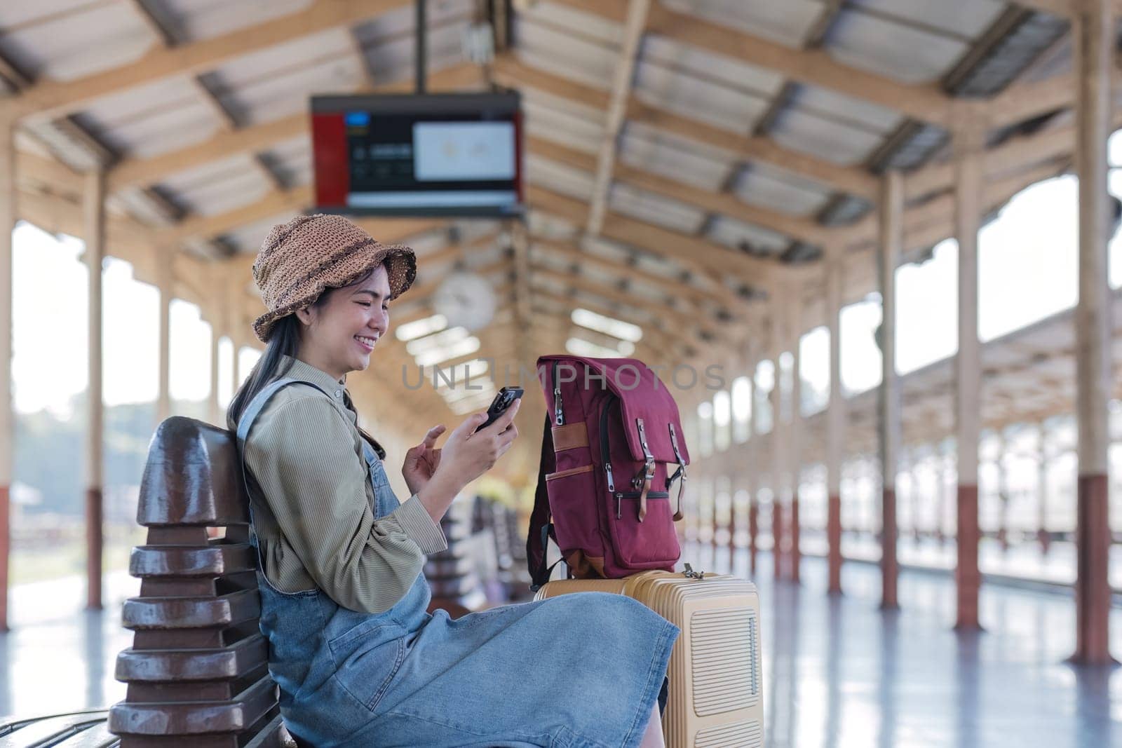 Young Asian woman in modern train station Female backpacker passenger sitting on a bench using a phone while waiting for a train at the train station to go on holiday. by wichayada
