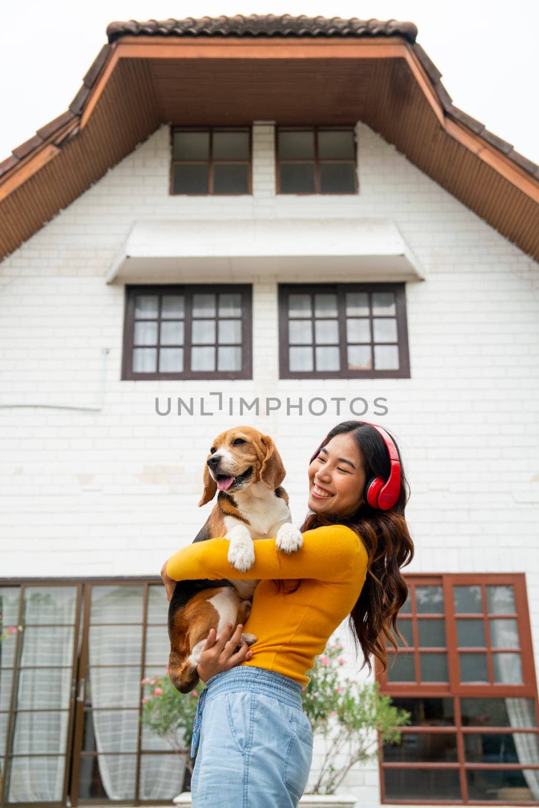 Vertical image of young Asian girl with red earphone hold and hug beagle dog and stand in front of her house and she look happy with her pet.