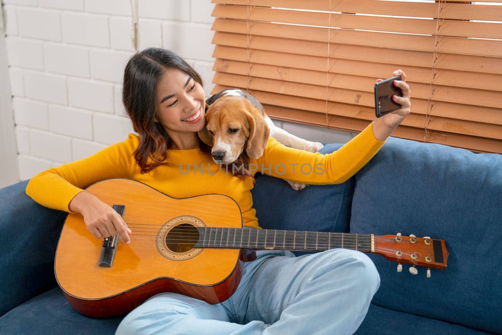 Young Asian girl enjoy to selfie with her dog during relax after play guitar on sofa in her house with day light.