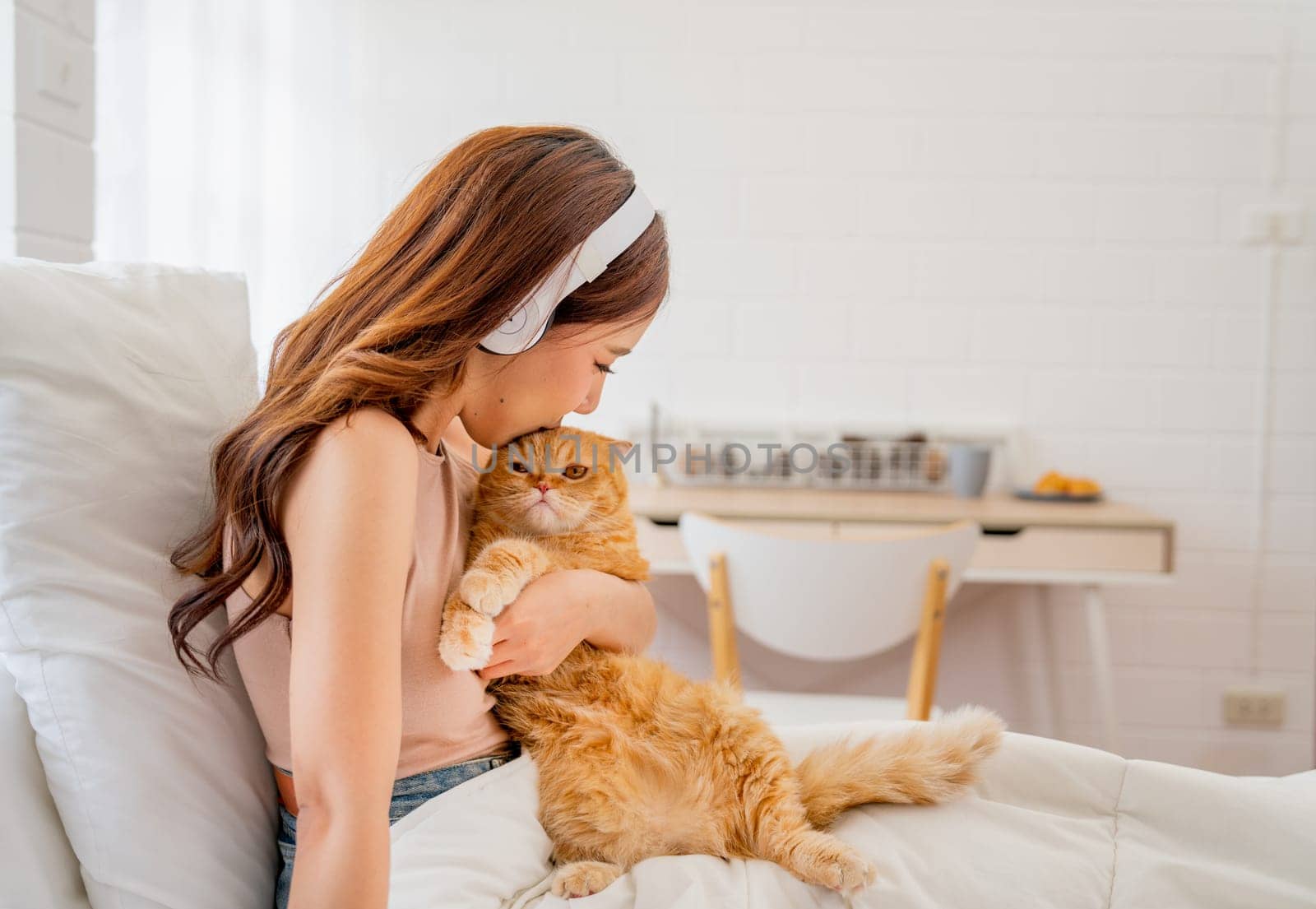 Pretty Asian woman with headphone hold and kiss orange cat sit on the bed in bedroom of her house.