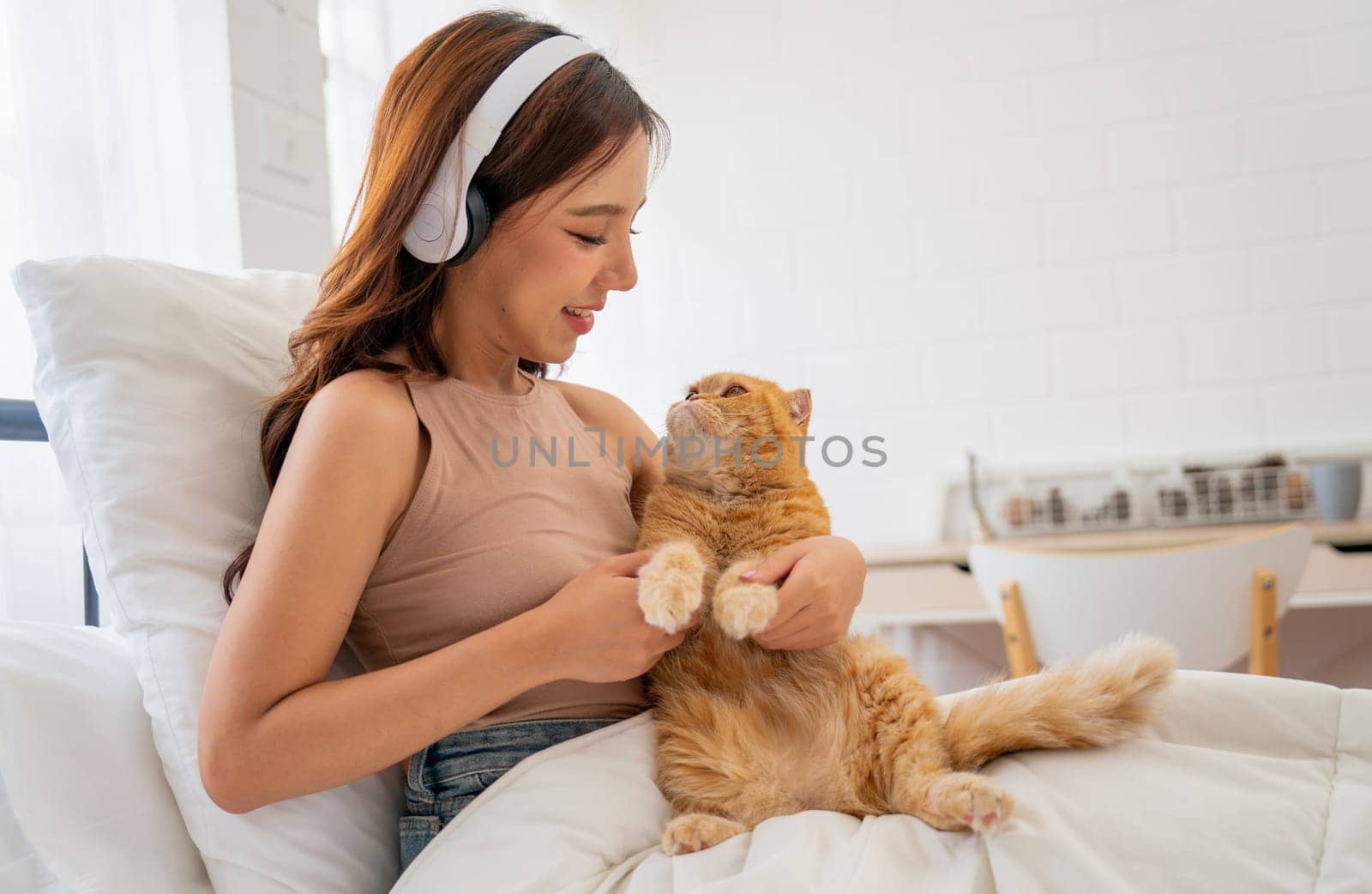 Pretty Asian woman with headphone hold and hug orange cat sit on the bed in bedroom with smiling and cat look at woman face.