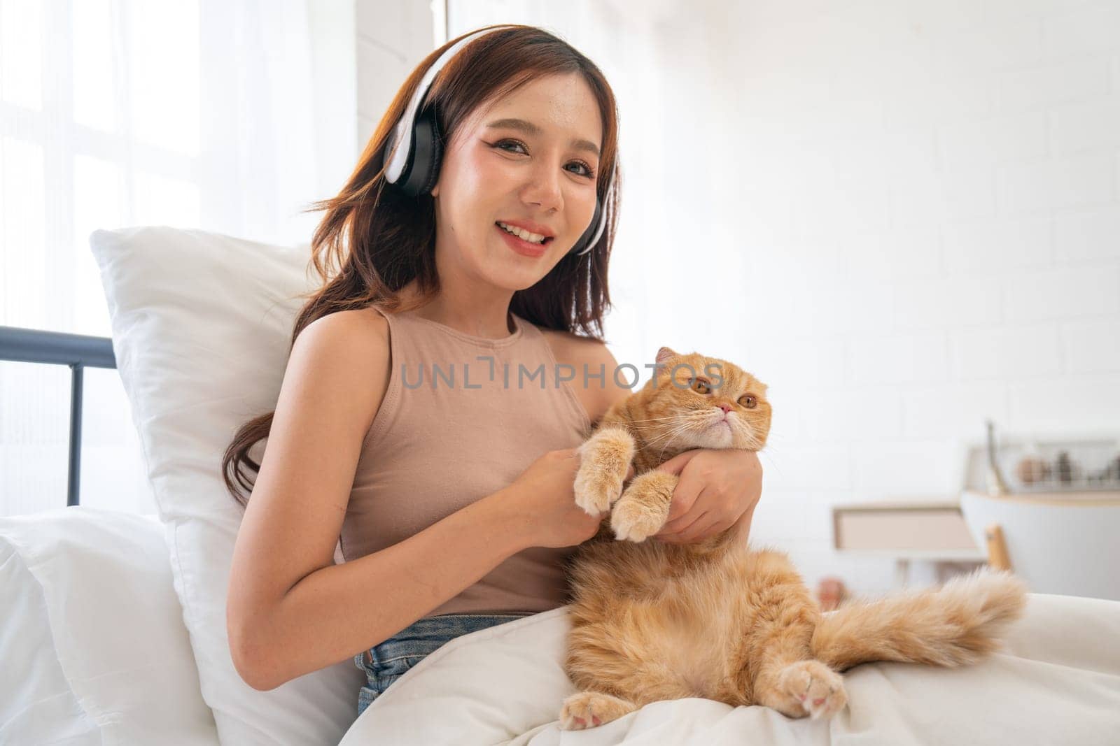 Lovely Asian girl with headphone hold and cudding orange cat and sit on bed also look at camera with smiling and day light. by nrradmin