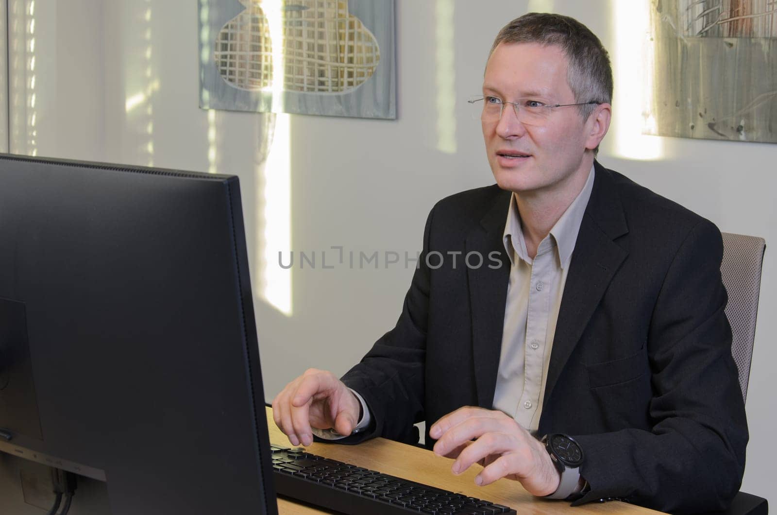 man works at computer in IT office. Middle-aged man in glasses and jacket. monitor in foreground. screen is not visible. On wall behind him glare of sun.