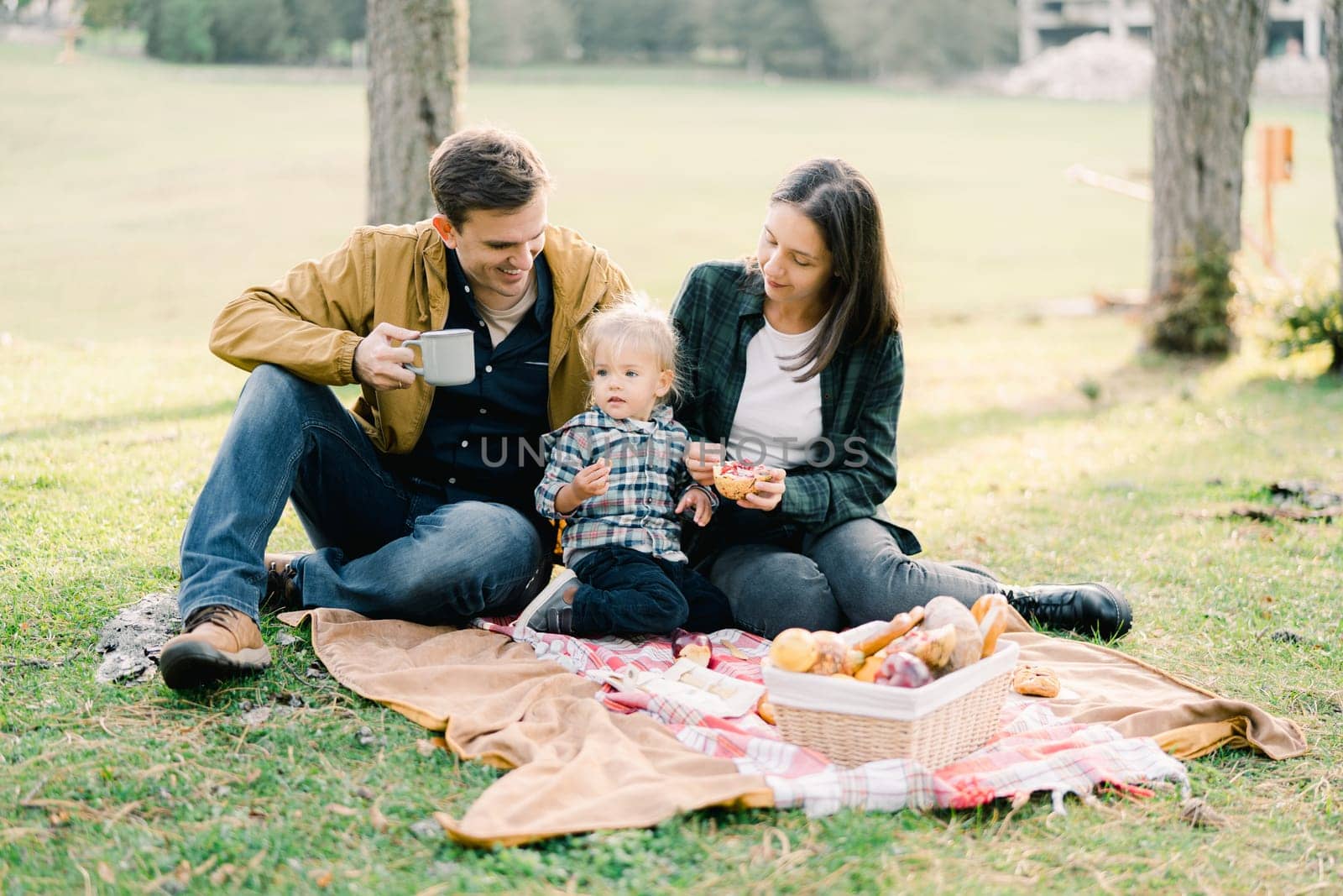 Dad with a mug in his hand sits near mom treating a little girl with a bun in the park. High quality photo