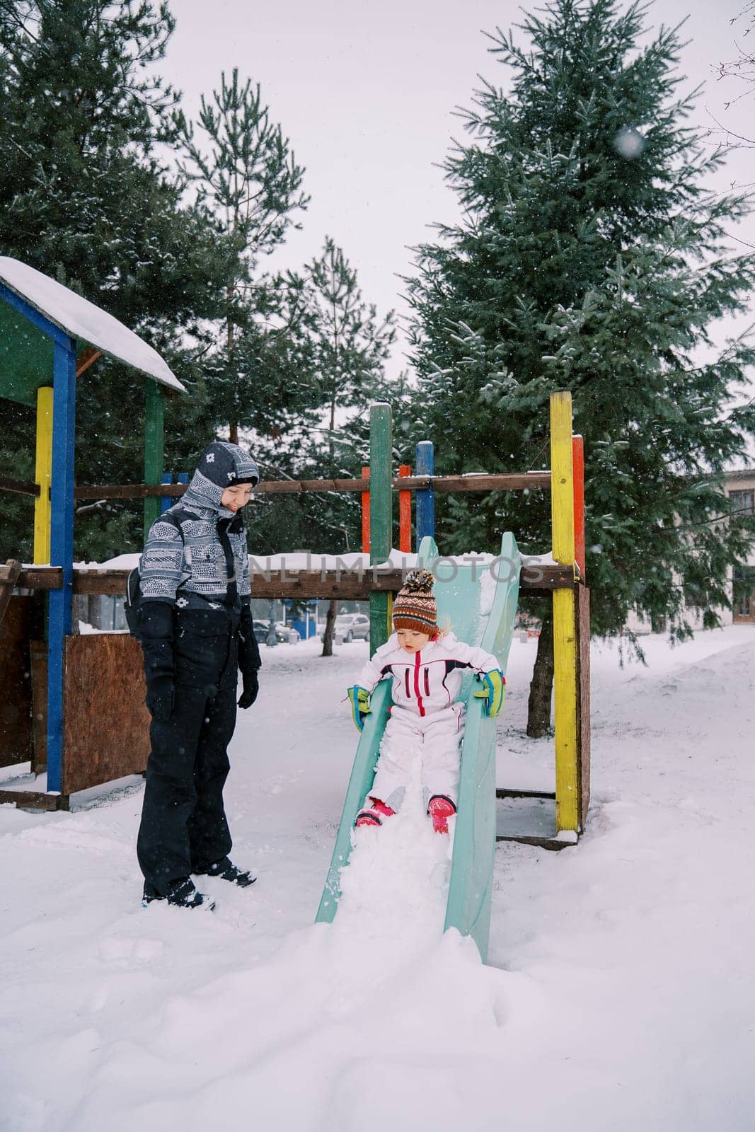 Mom looks at a little girl sliding down a snow-covered colorful slide by Nadtochiy