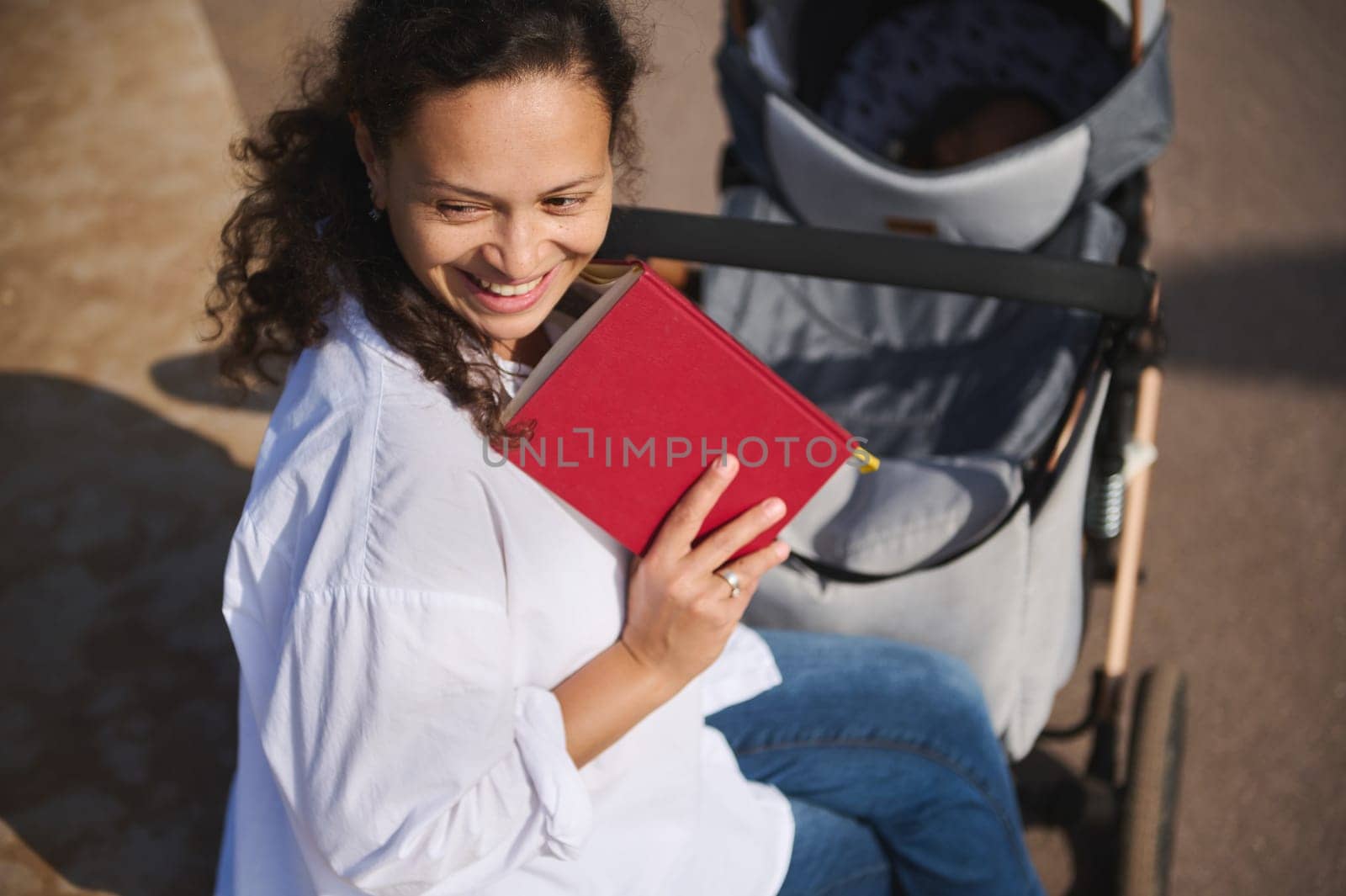 Beautiful Latin woman smiling looking aside, holding book and pushing baby pram with her sleeping child. Maternity leave by artgf