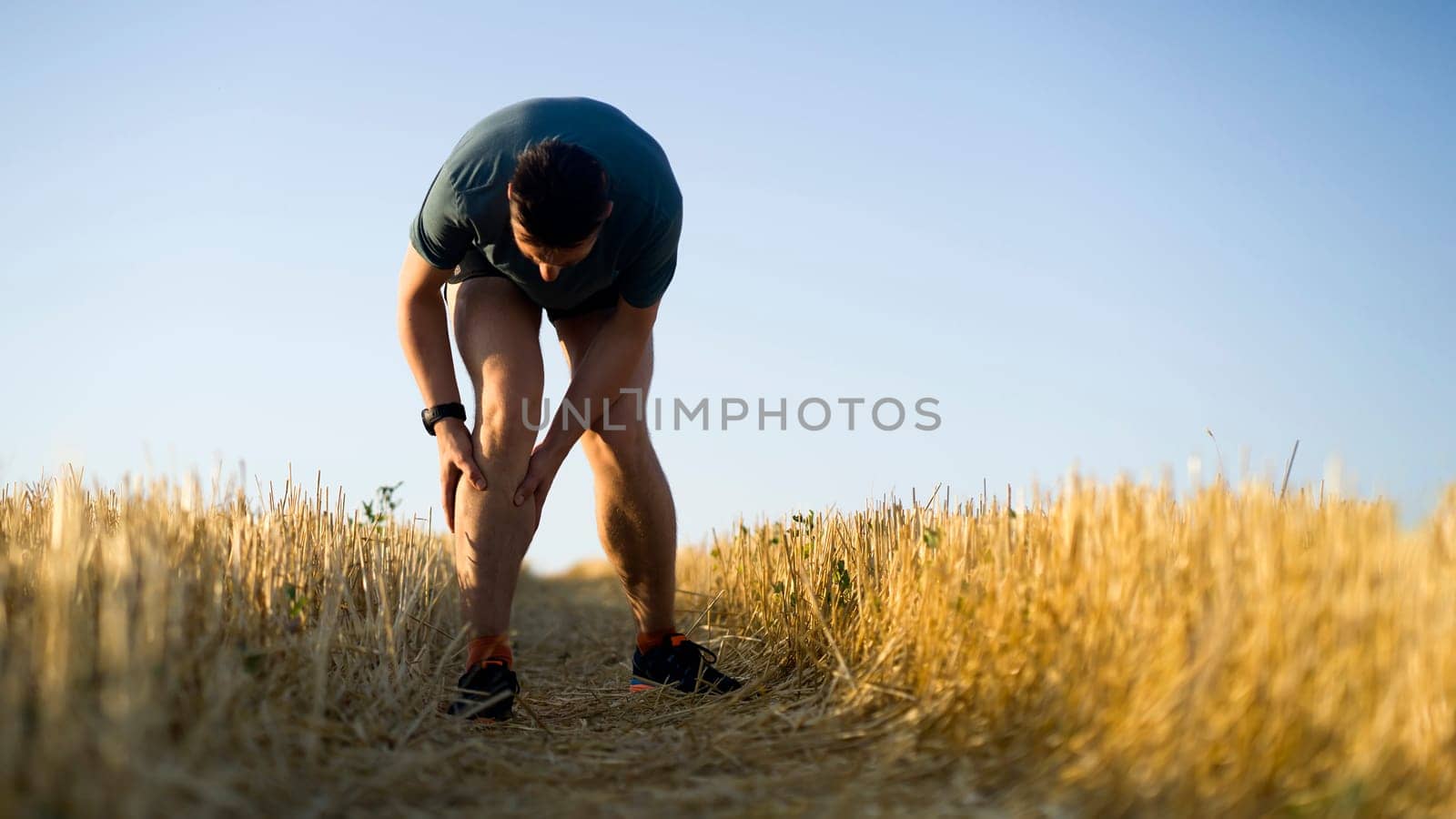 A young man jogging at sunset on the trail across the field, the tired athlete holds on to his sore knee.