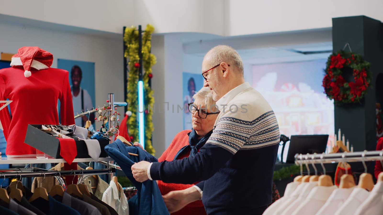 Senior customer browsing through xmas decorated clothing store racks with husband in Christmas shopping spree. Aged couple searching for stylish blazer as present for son during winter holiday season