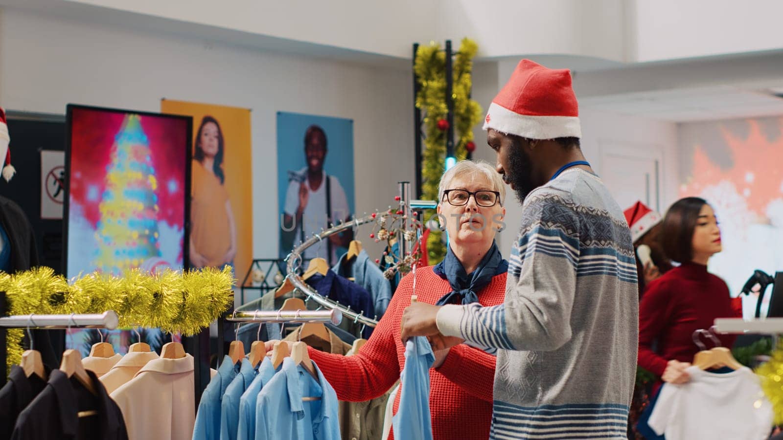 African american worker wearing Santa hat helping woman browsing for clothes in festive Christmas decorated clothing store. Employee assisting elderly customer in festive adorn fashion shop