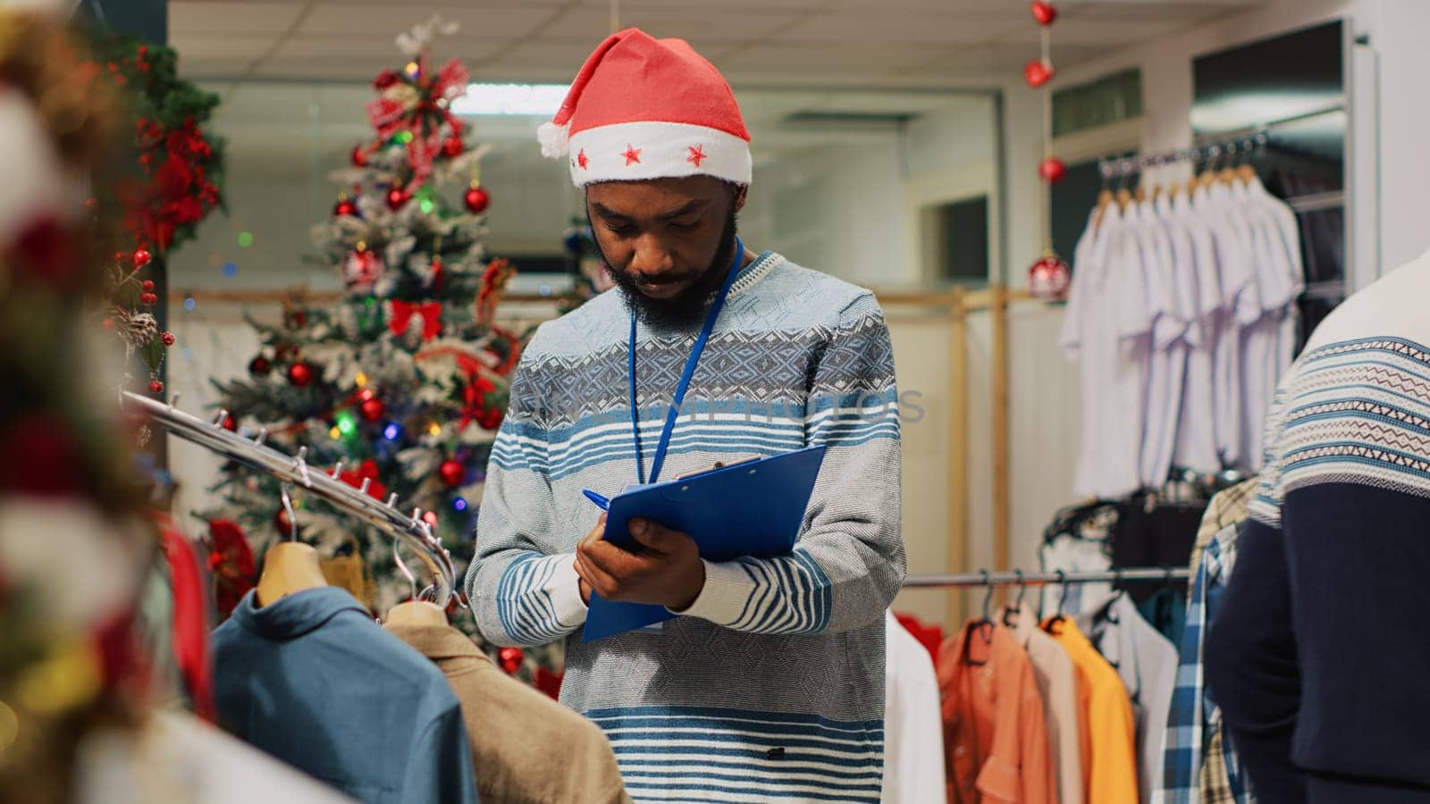 African american employee browsing through clothing racks in Christmas decorated fashion store, writing down adjusted prices on clipboard. Worker inspecting blazers, looking for damages