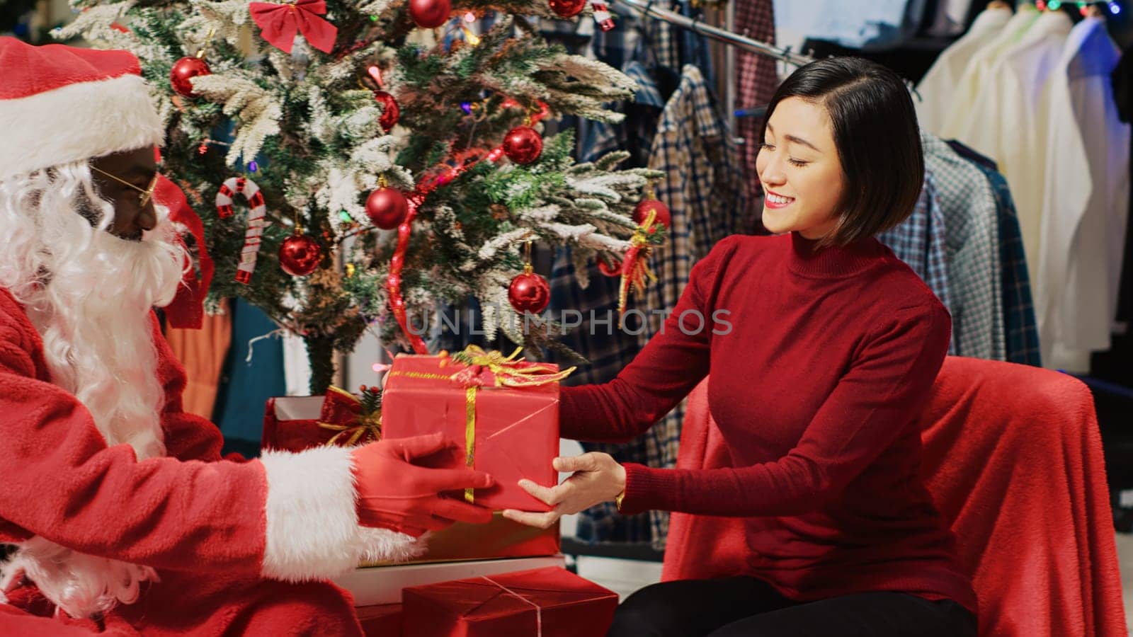 Customer in fashion store talking with actor dressed as Santa Claus, sitting next to Christmas tree during festive promotional event. Client receiving present from worker during holiday season