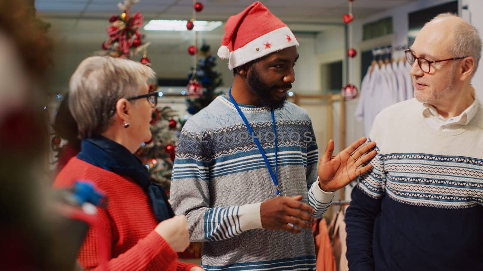 Retail assistant in festive decorated clothing store helping elderly couple during their Christmas frenzy shopping spree. Employee offering advice to senior customers during winter holiday season