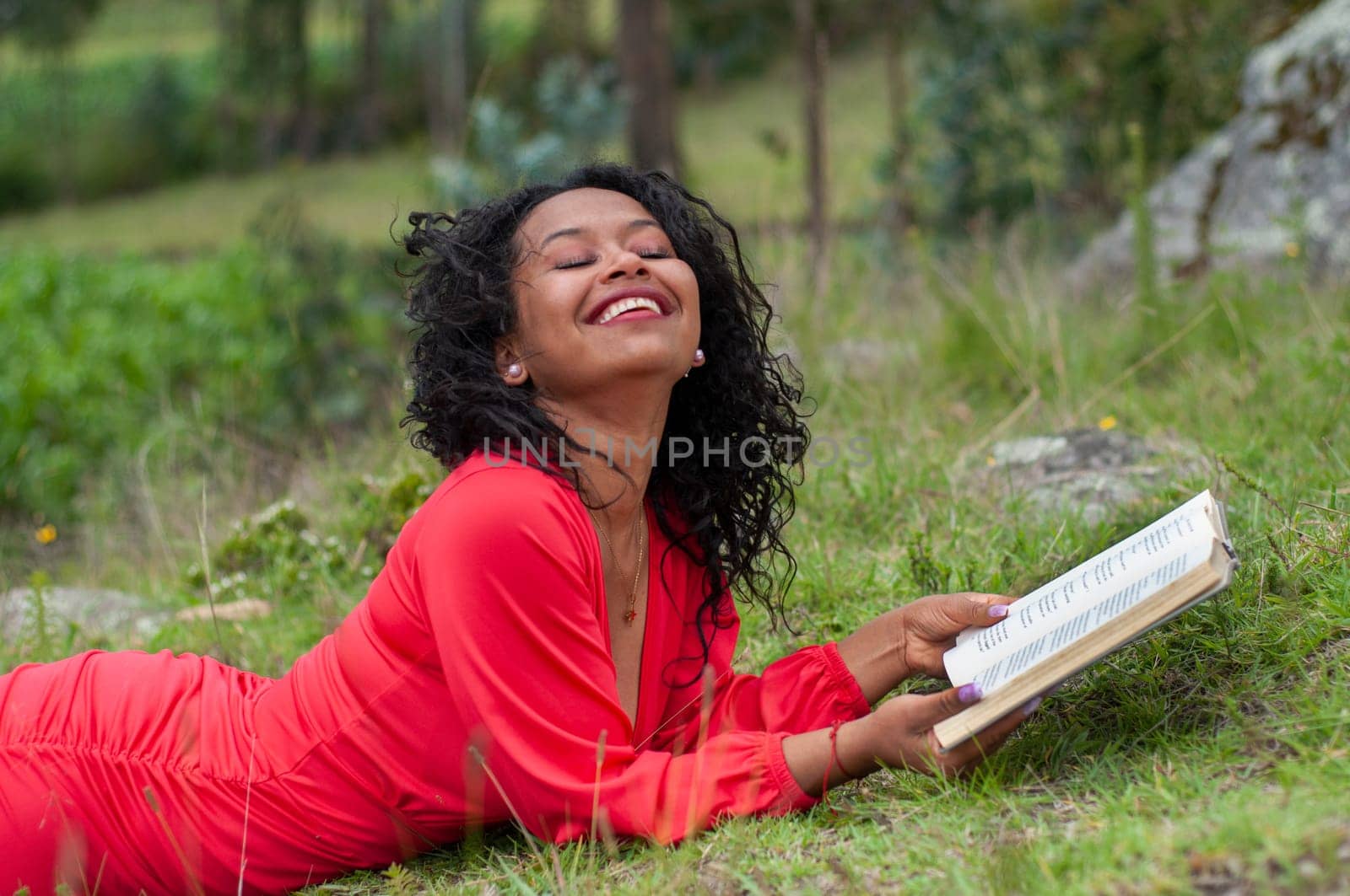 young latin girl lying on the grass of a mountain with a book in her hands, very happy with her head up, eyes closed and smiling. book day by Raulmartin