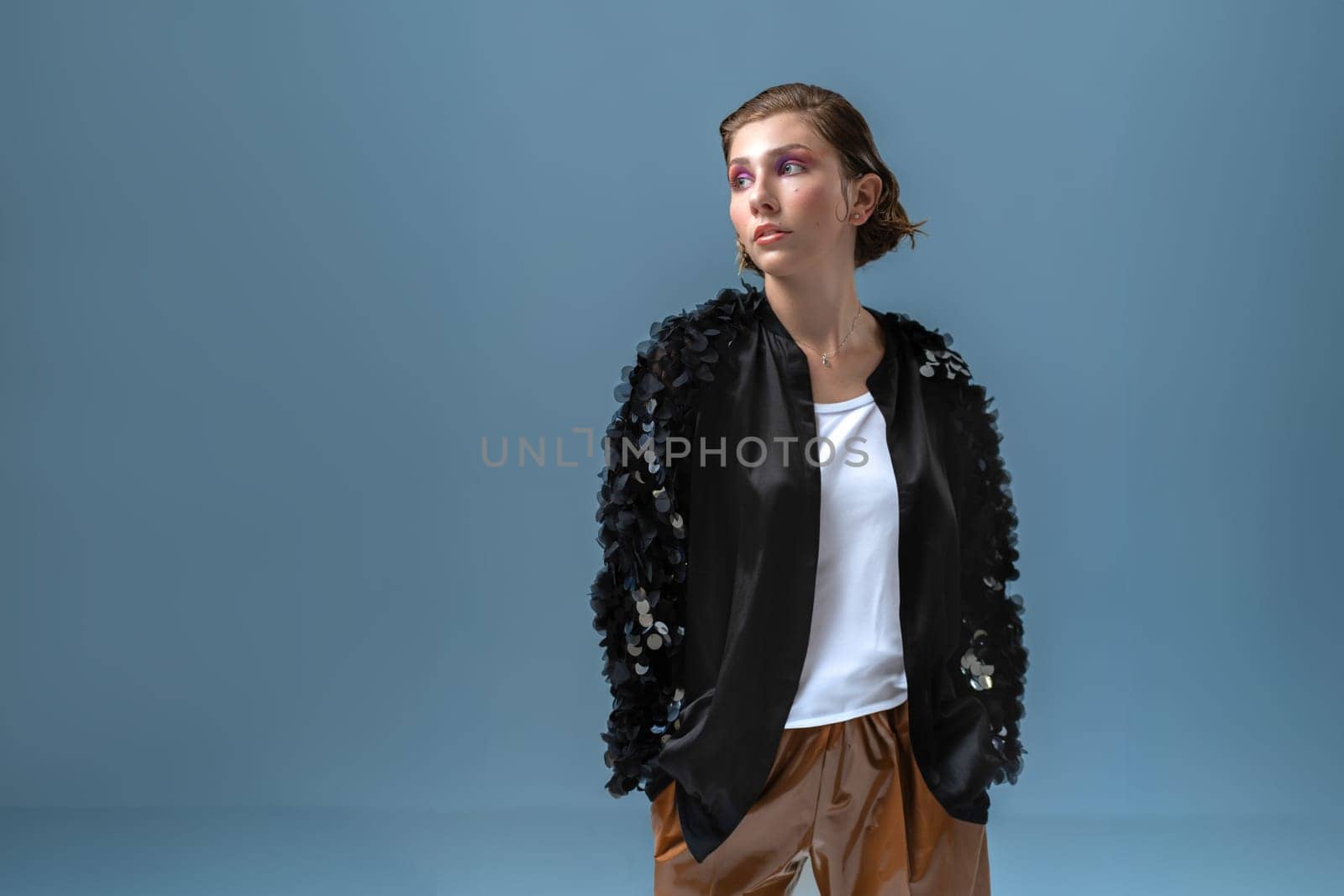 Fashionable confident woman wearing stylish black jacket, standing hands in pocket isolated on blue studio background looking away, middle shot. Studio fashion portrait