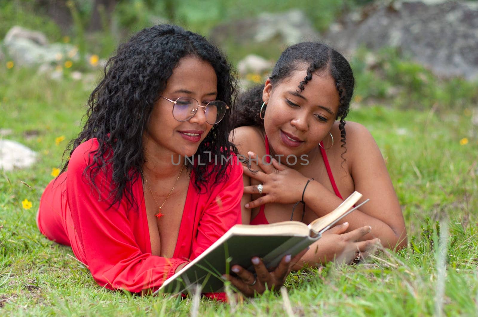 two latina friends lying face down together reading a book attentively the girl holding the book has a pair of glasses. book day by Raulmartin