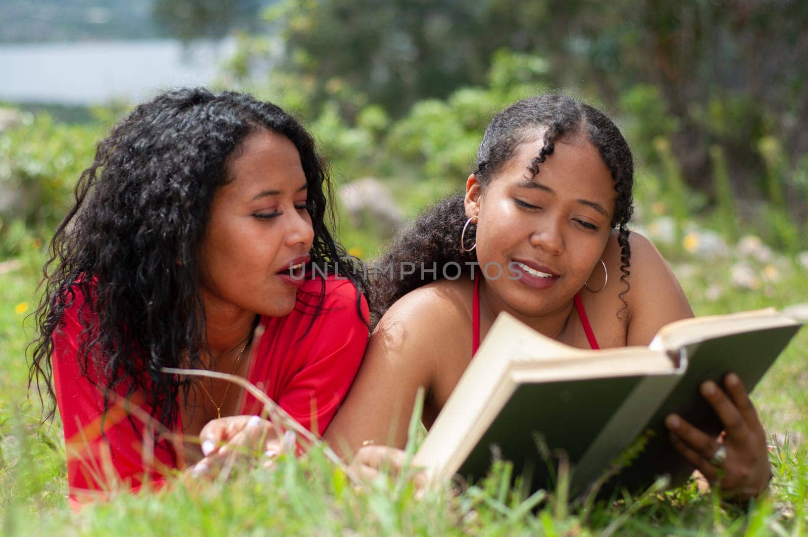 two young women from colombia with black curly hair reading a book lying on a mountain on a very sunny day. book day. by Raulmartin