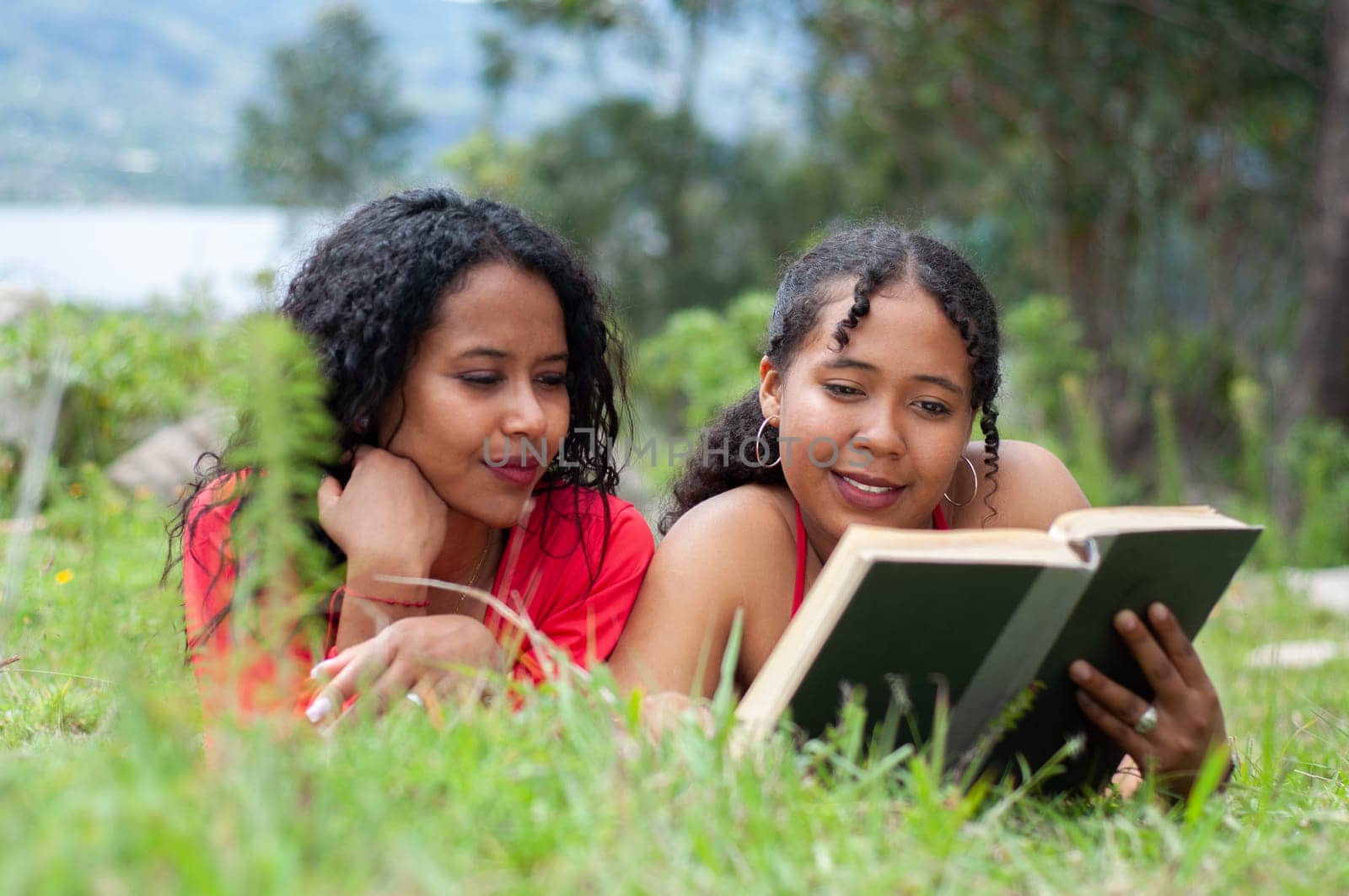 two latin sisters reading a book in a peaceful and quiet environment surrounded by trees and nature near a lake. book day. by Raulmartin