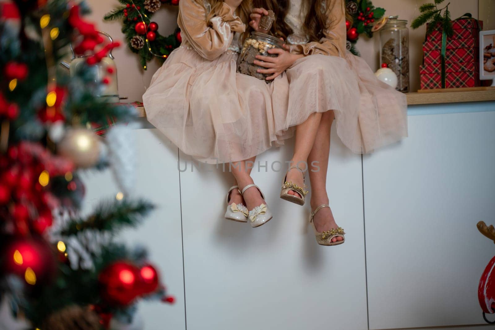 Two cute little girls in beige dresses are sitting on the table holding cookies. New Year decoration. Luxurious New Year's interior