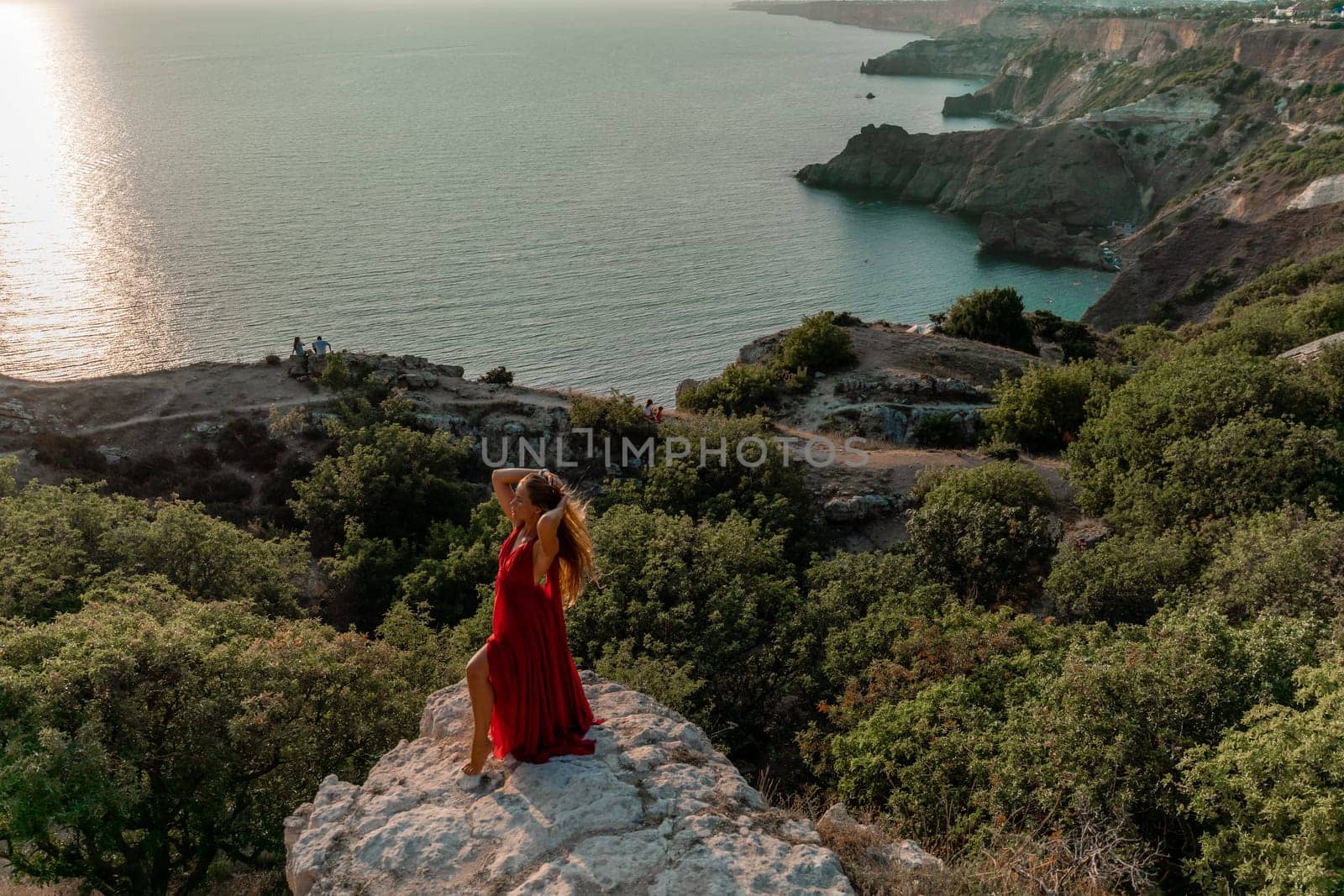 Woman sunset sea red dress, side view a happy beautiful sensual woman in a red long dress posing on a rock high above the sea on sunset