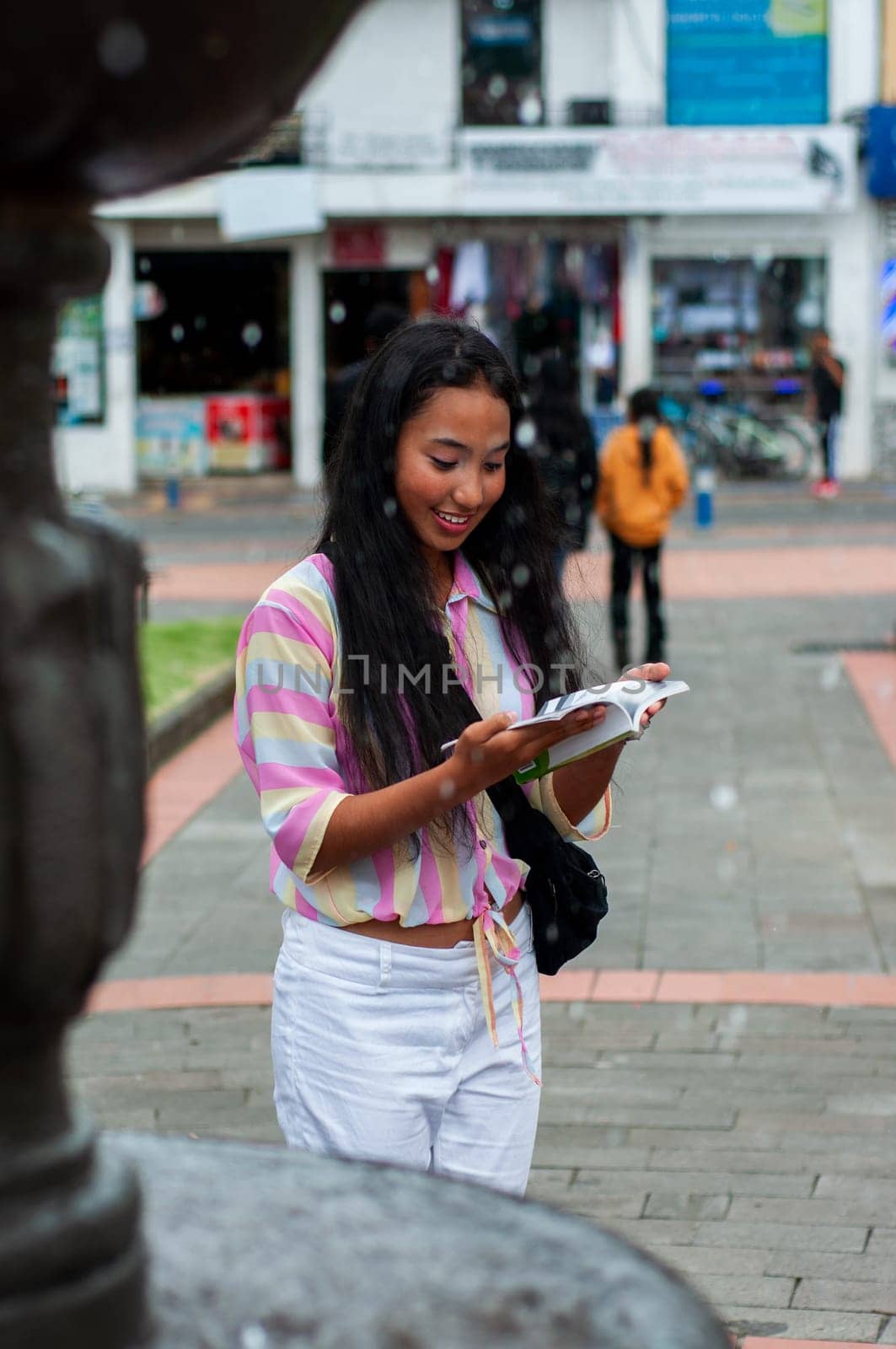 pretty teenage girl from ecuador standing next to a fountain reading a book while spending a nice relaxing afternoon. book day by Raulmartin
