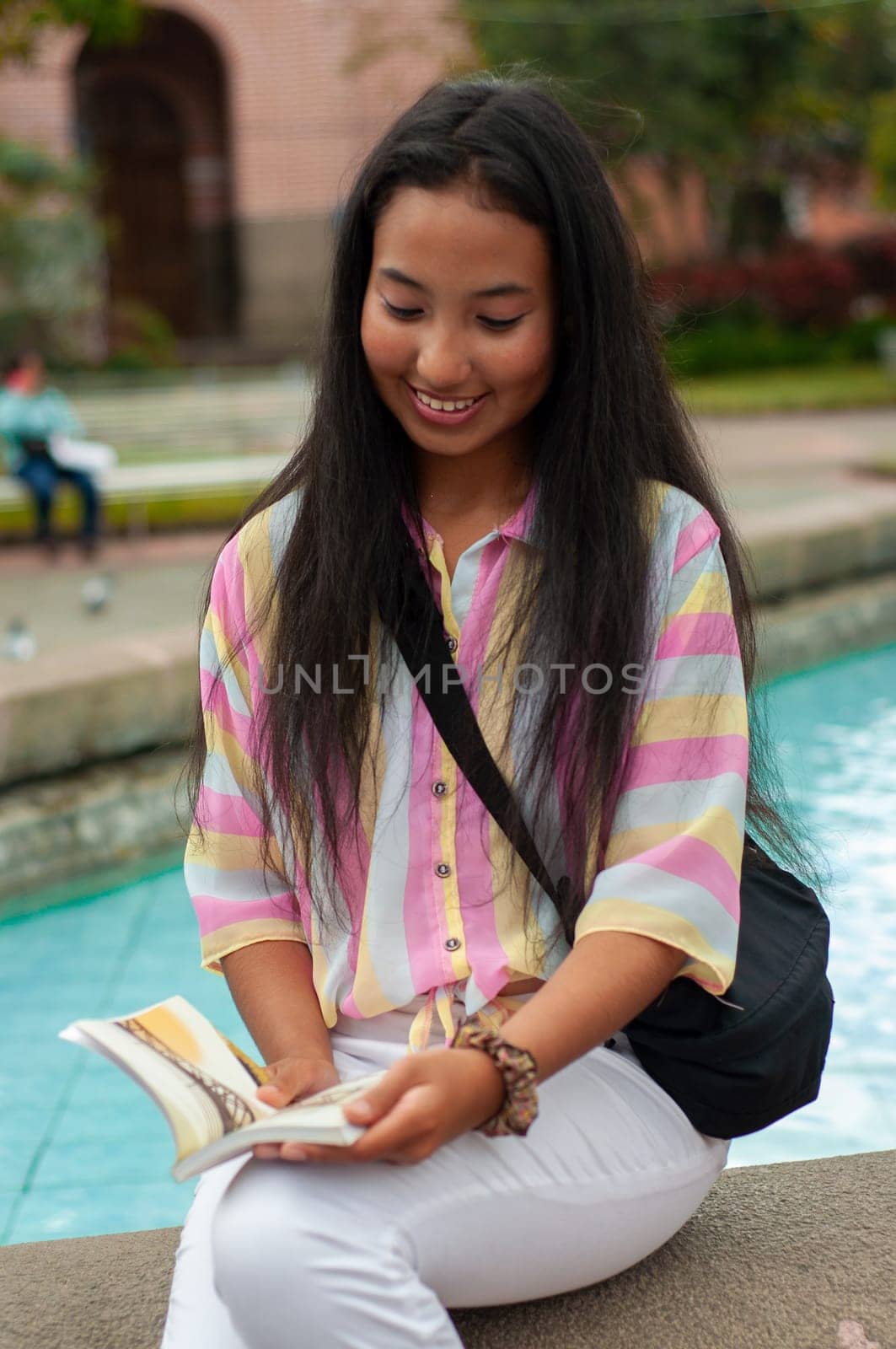 young girl from latin america sitting at the edge of a public fountain filled with water reading a book while smiling and enjoying her reading. book day by Raulmartin