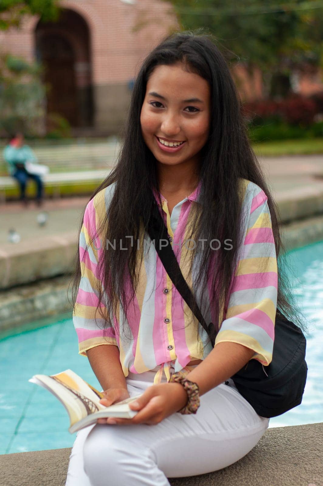 young woman from south america sitting in a fountain with long flowing hair smiling and looking at the camera while holding a paper book open in the middle. book day. by Raulmartin