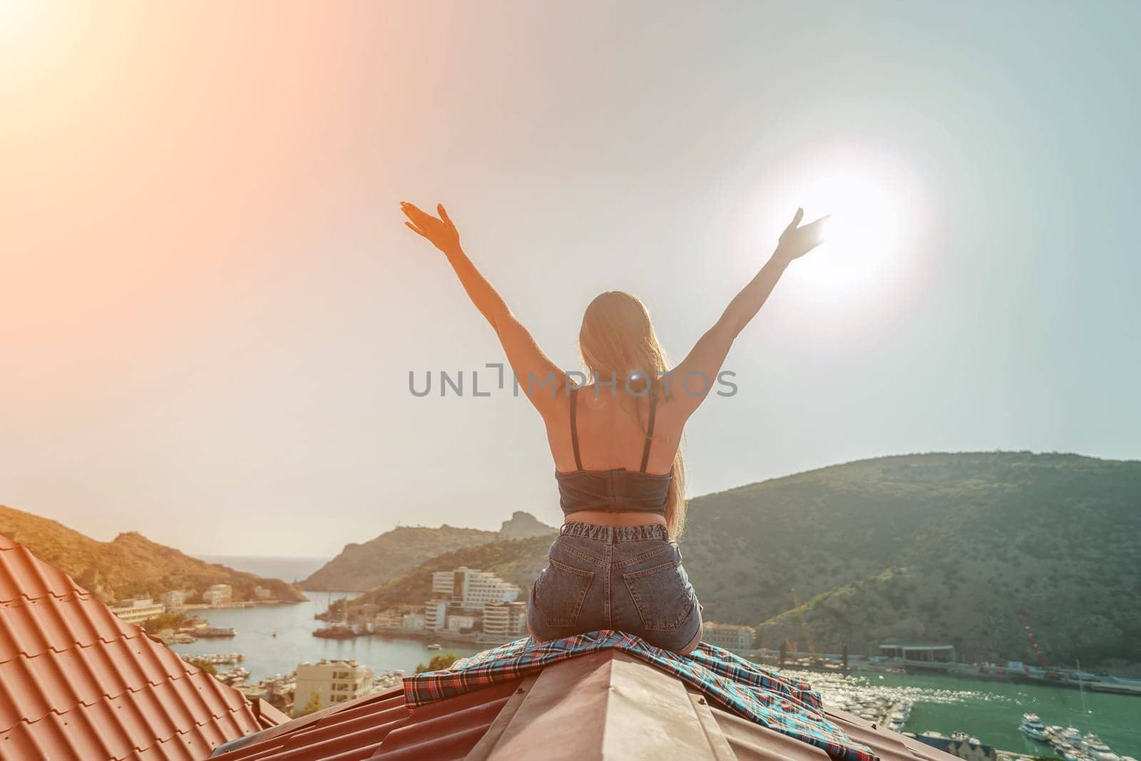 Woman sits on rooftop with outstretched arms, enjoys town view and sea mountains. Peaceful rooftop relaxation. Below her, there is a town with several boats visible in the water.