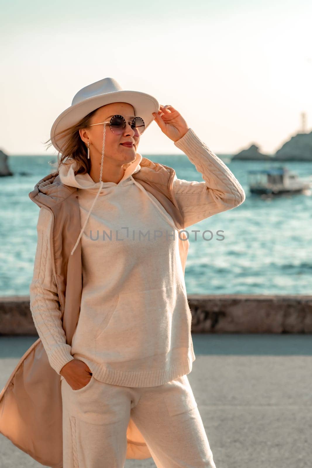 Happy blonde woman in a white suit and hat posing at the camera against the backdrop of the sea.