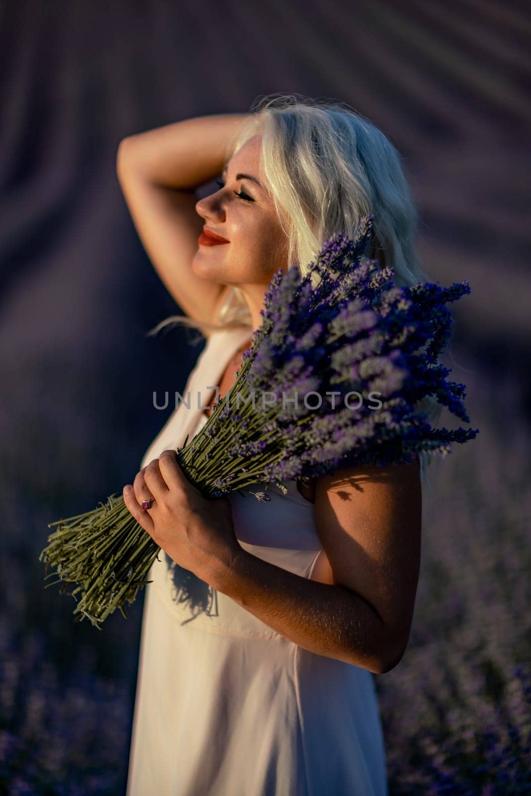 Blonde woman poses in lavender field at sunset. Happy woman in white dress holds lavender bouquet. Aromatherapy concept, lavender oil, photo session in lavender.